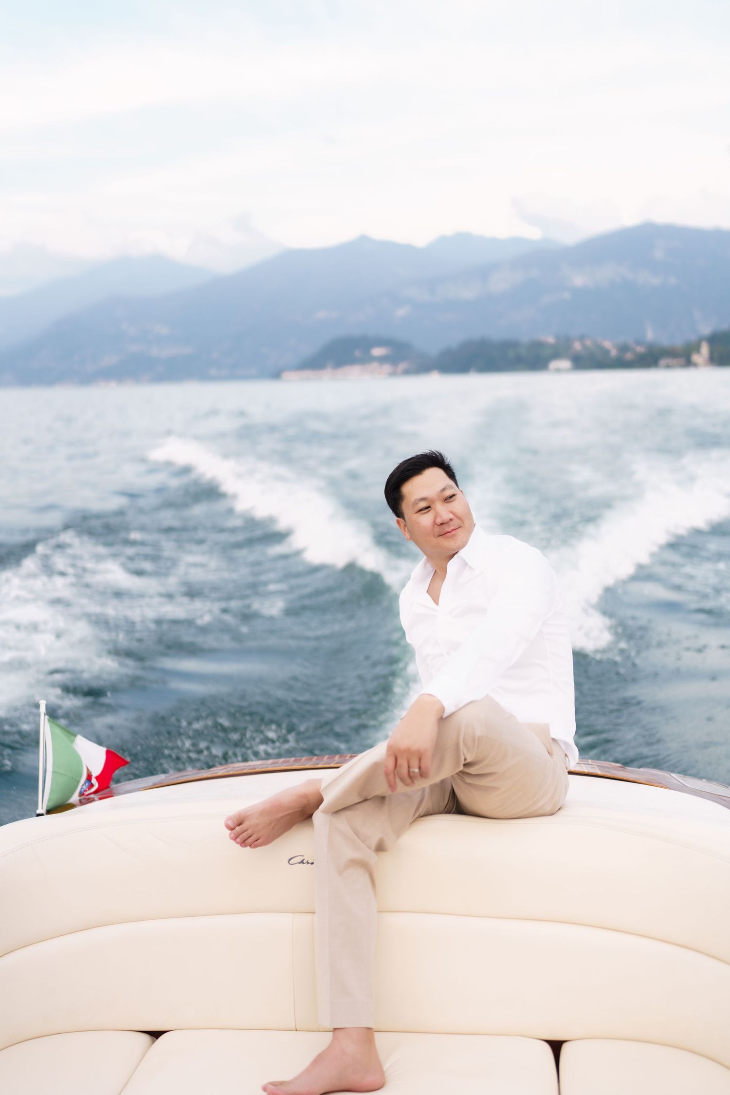 Person relaxes on white cushions of a luxury yacht while cruising past misty mountains on calm waters.