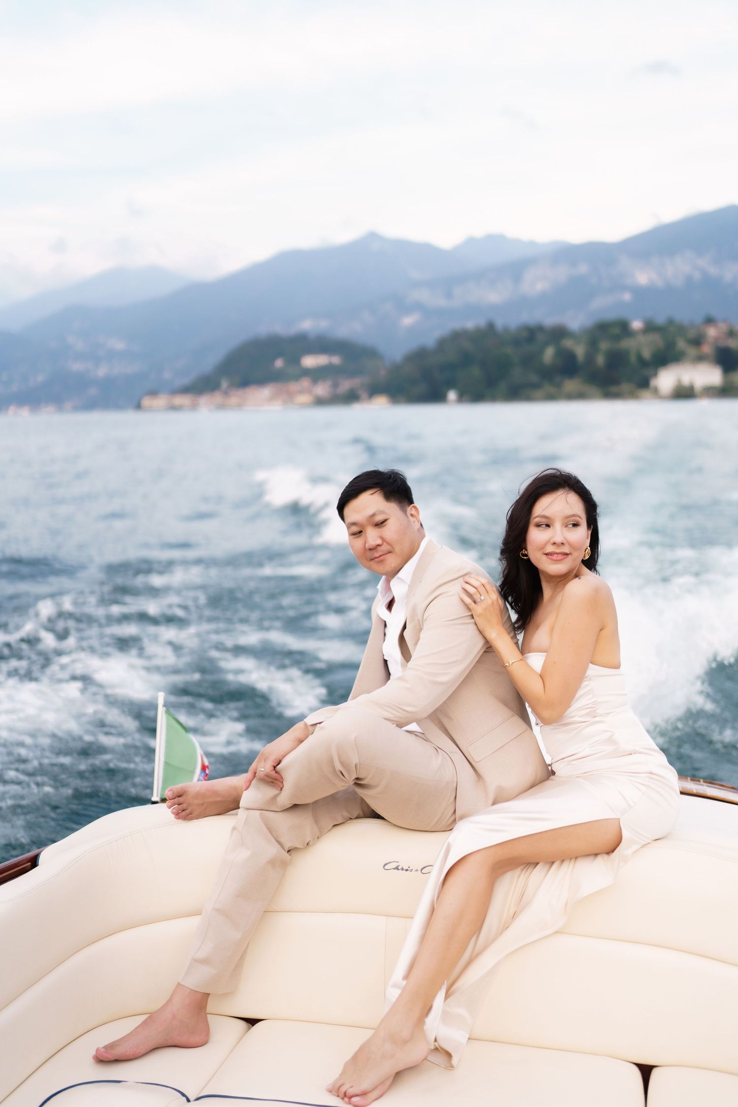 A couple relaxes together on a white boat cushion with mountains and water visible in the background.