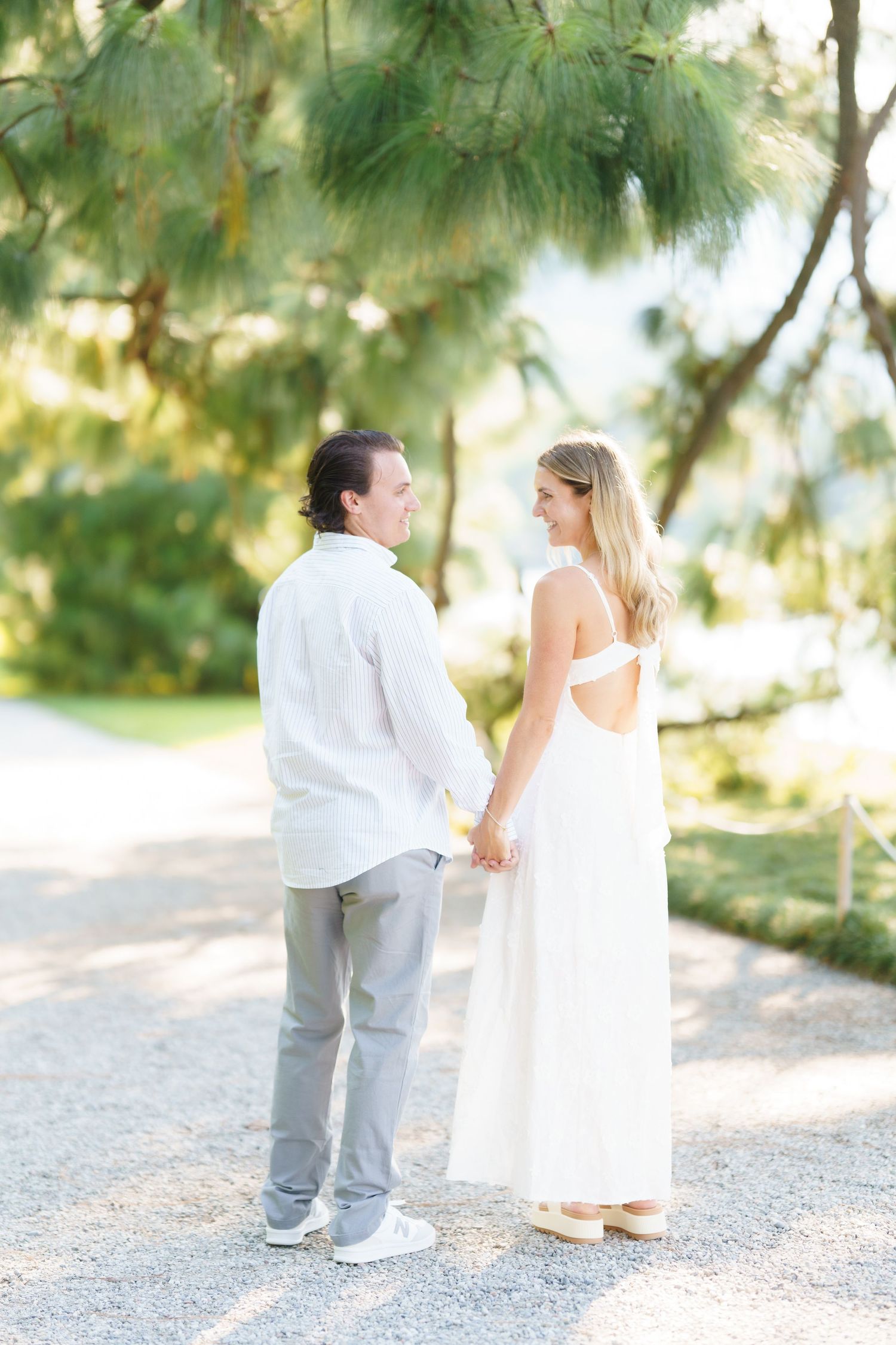 A couple in white and gray attire walks along a gravel path surrounded by green pine trees in a romantic outdoor setting.