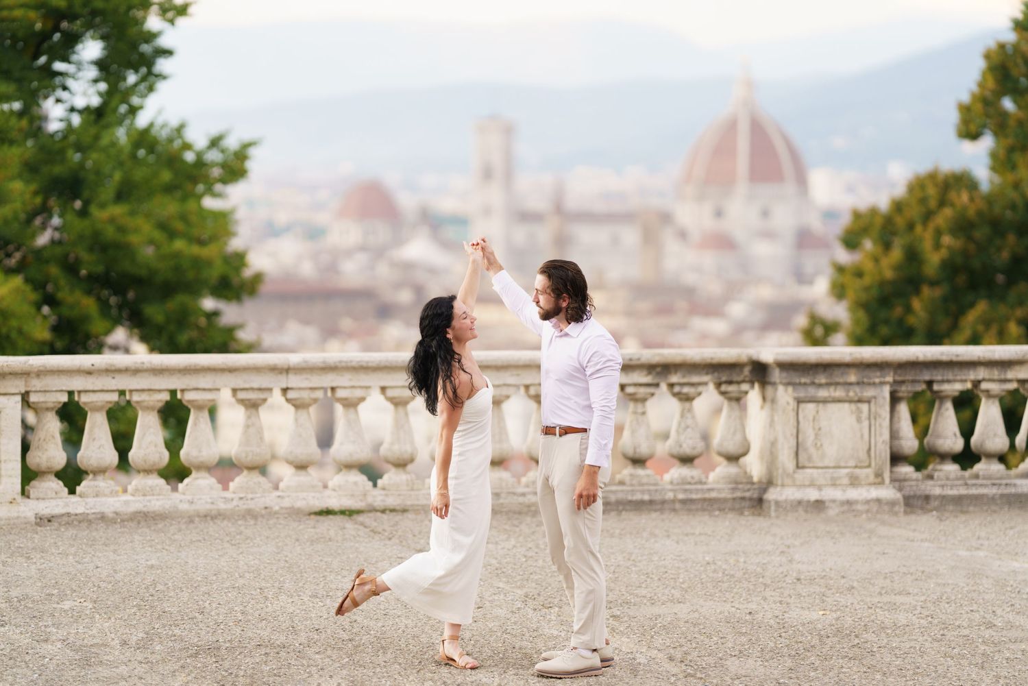 Two people in white outfits enjoy a romantic moment at a scenic overlook with Florence's Duomo cathedral in the background.