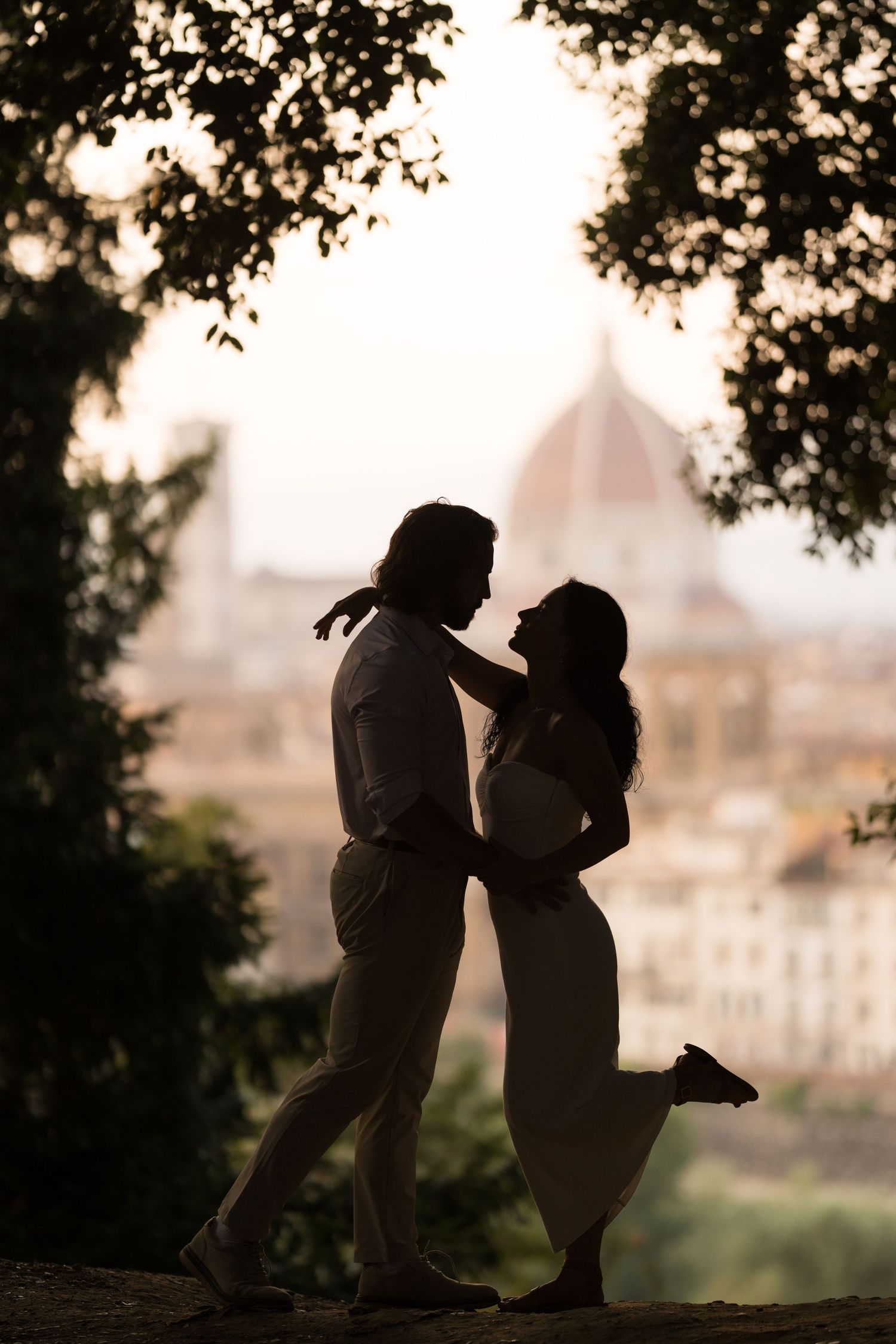 Silhouettes of two figures share a tender moment at sunset with a city skyline in the background.