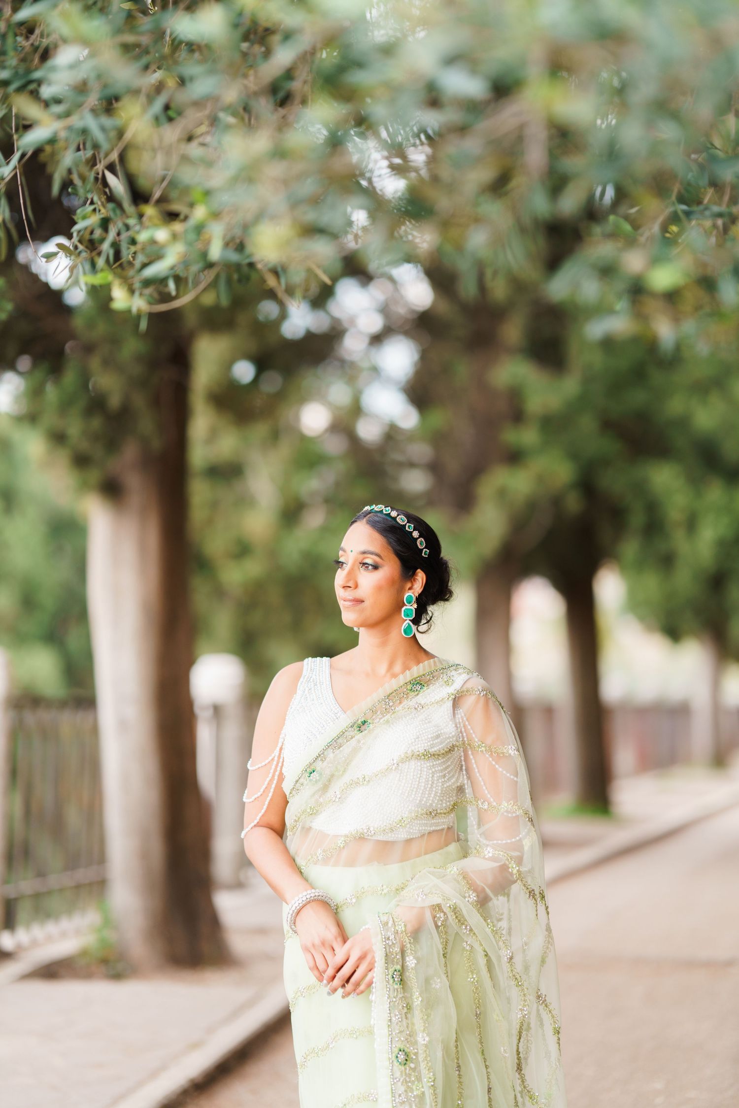 A bride in an elegant cream-colored sari poses gracefully in a tree-lined walkway during an outdoor photo session.
