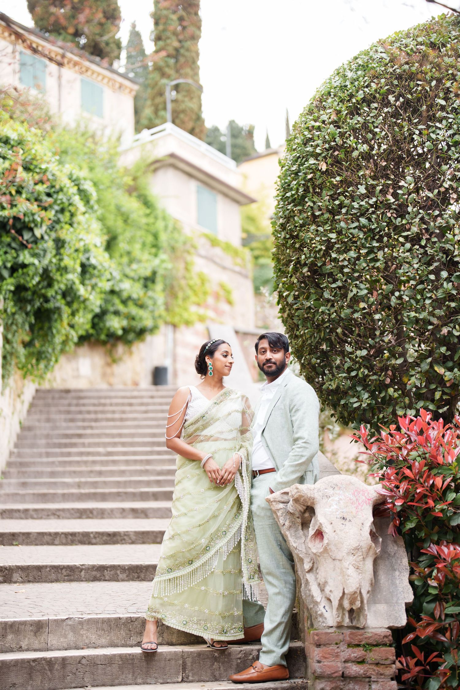 photographer verona A couple in traditional attire sits on stone steps surrounded by lush greenery and blooming flowers in a garden setting.