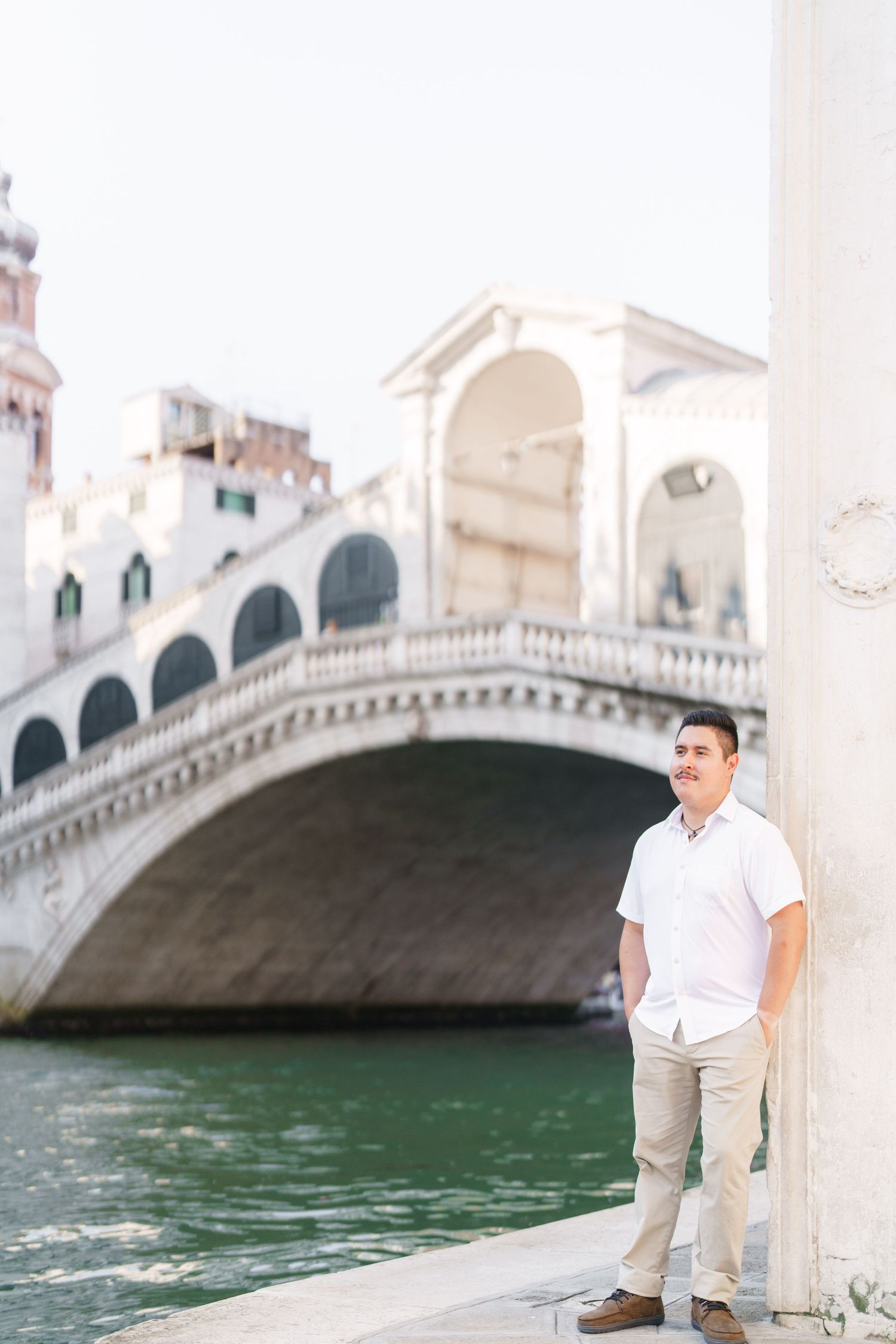 A romantic couple stands by a Venetian canal with an arched stone bridge in the background.
