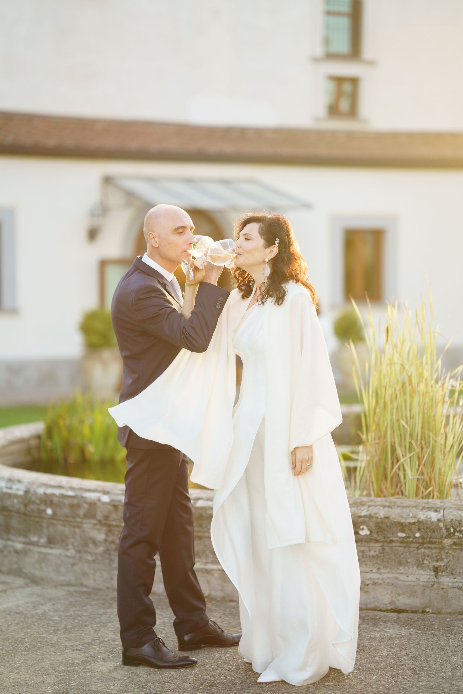 A romantic wedding moment captured as newlyweds toast with wine glasses against a sunlit backdrop.
