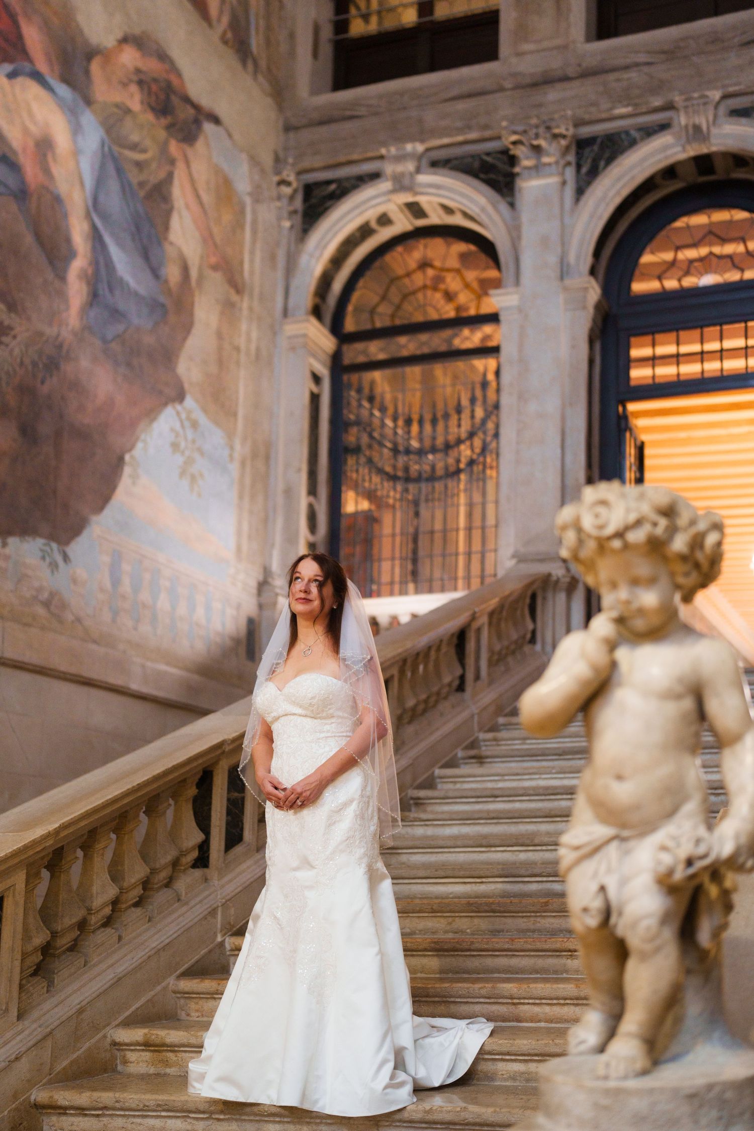Elegant bride in white dress poses on grand marble staircase with ornate cherub statue and frescoed walls.