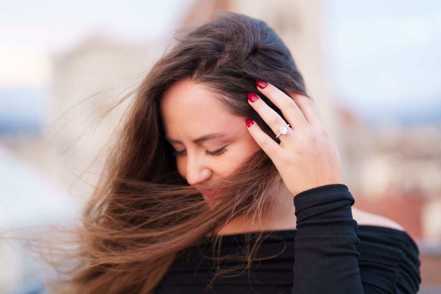 Woman in black sweater with windswept brown hair smiles with eyes closed against blurred outdoor background.