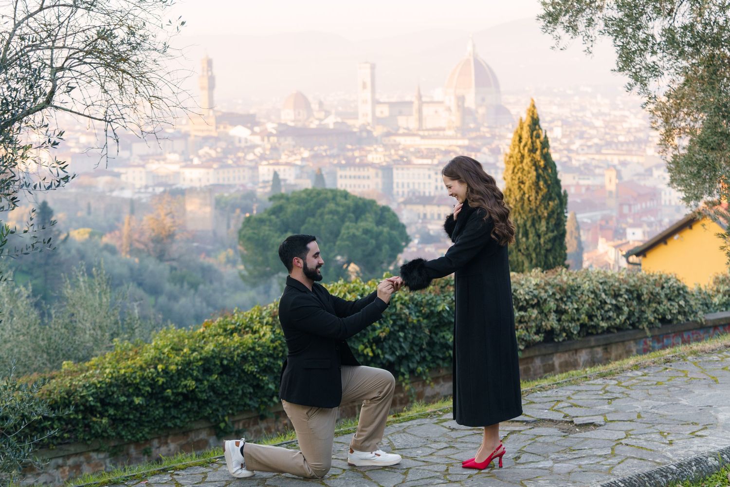 Two people sit on a bench overlooking a misty cityscape during sunset, creating a romantic silhouette.