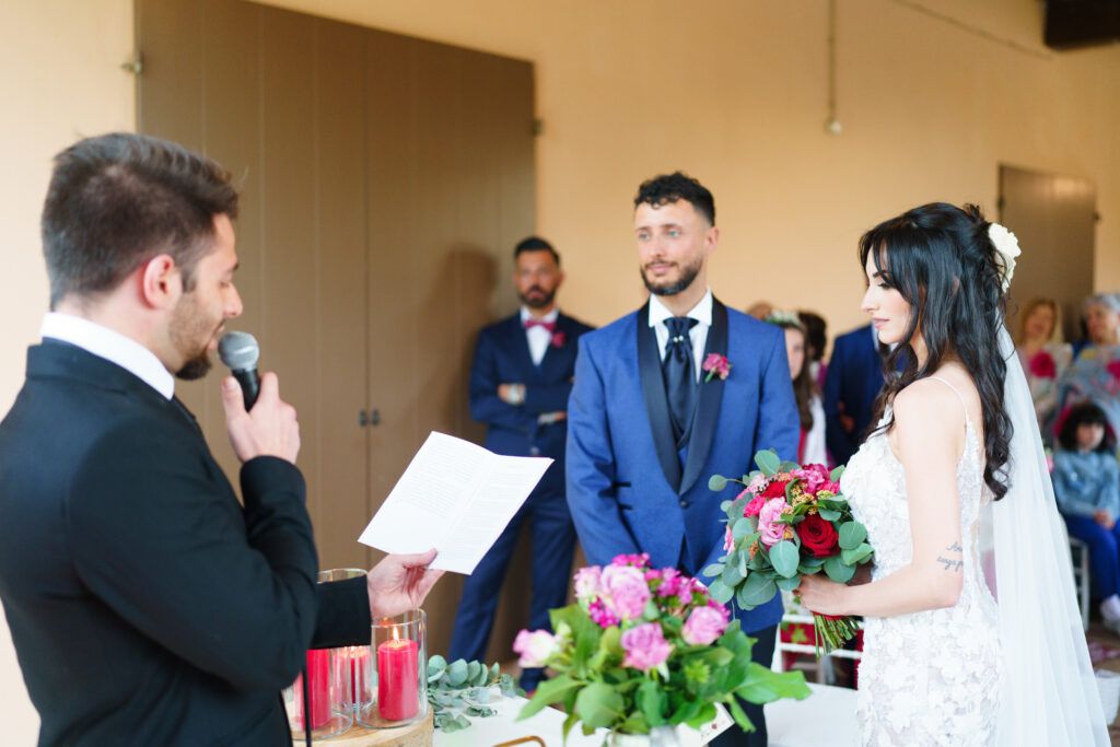 Wedding ceremony in progress with officiant reading from book as couple and guests gather in indoor venue.