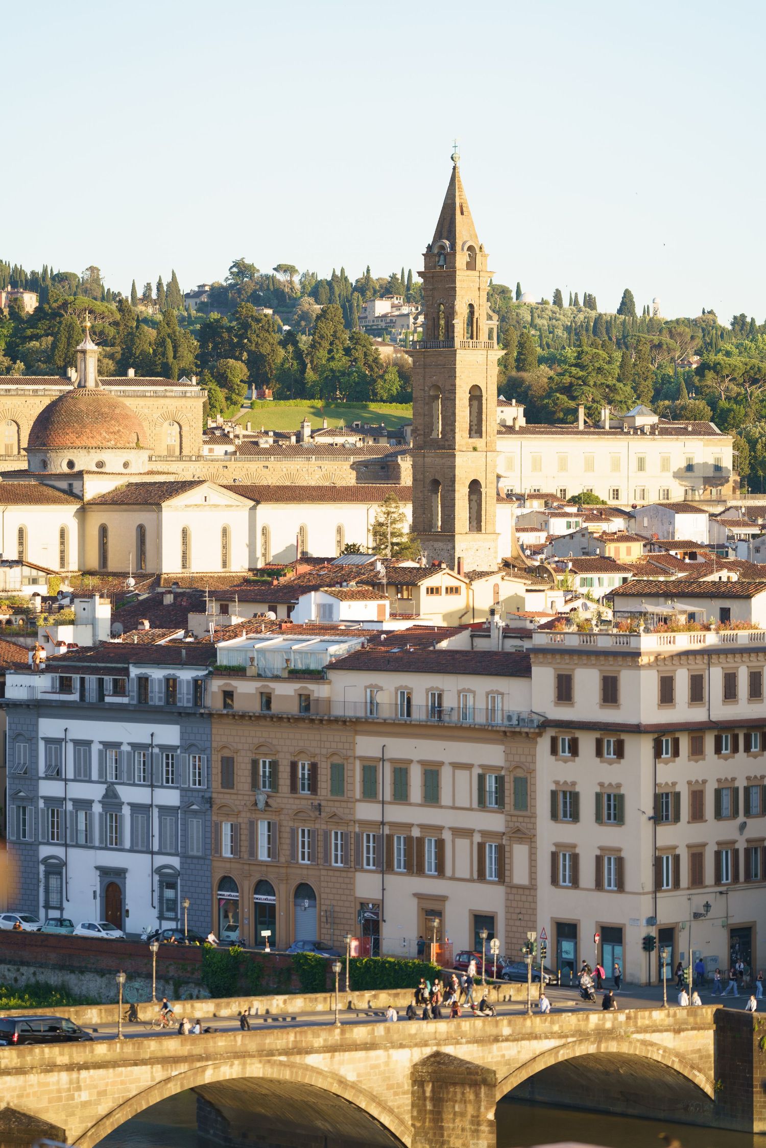 View of Florence's historic architecture with church tower and Arno River bridge in warm afternoon light.