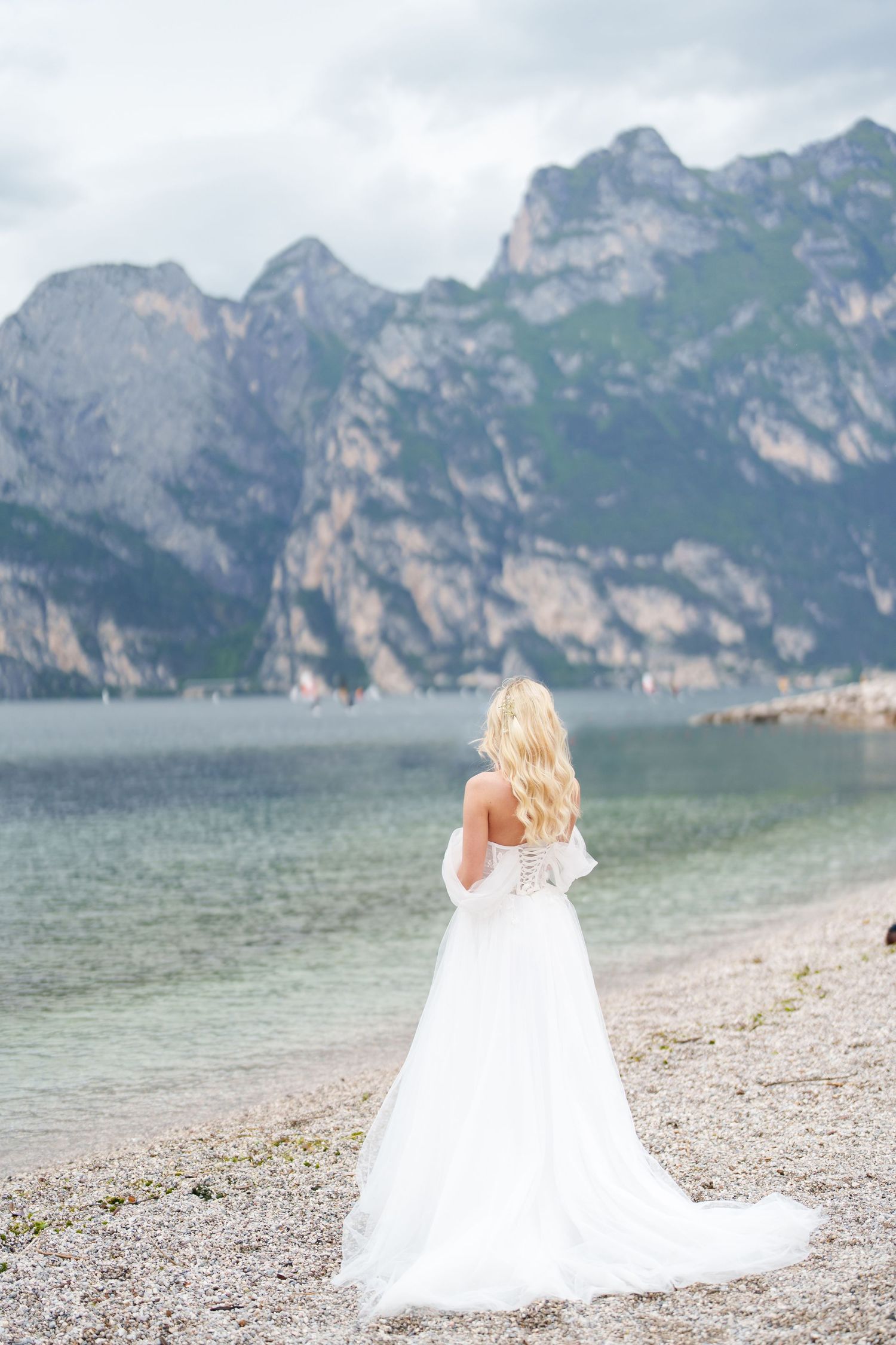 A bride in a flowing white gown stands on a rocky beach shore with dramatic mountain peaks rising above Lake Garda.