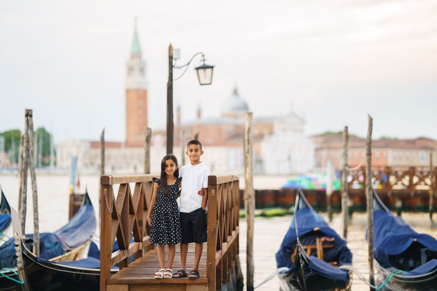 Two children pose on a wooden dock with gondolas and San Giorgio Maggiore island in Venice, Italy.
