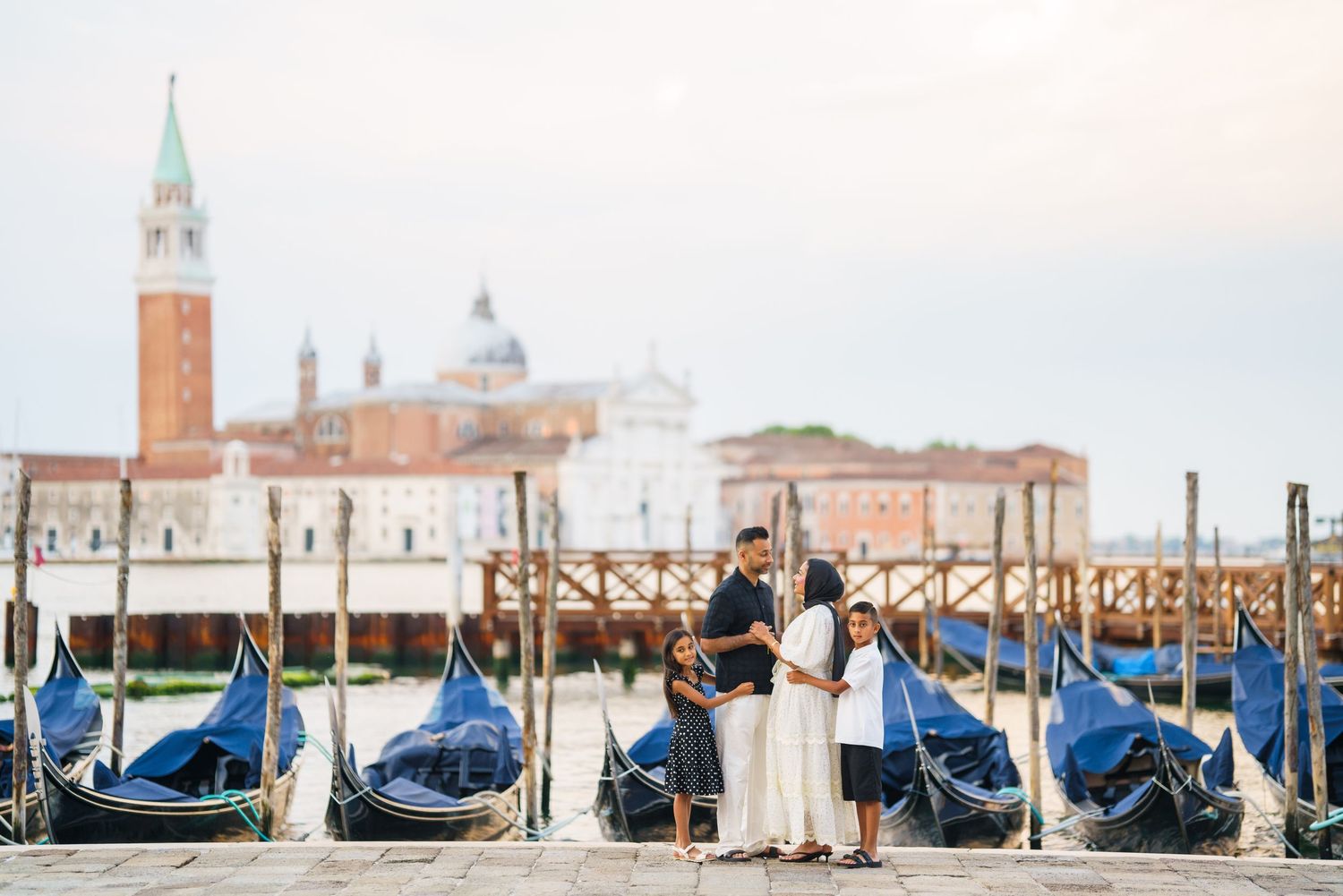 Family posing by gondolas in Venice with San Giorgio Maggiore island in the background at dusk.