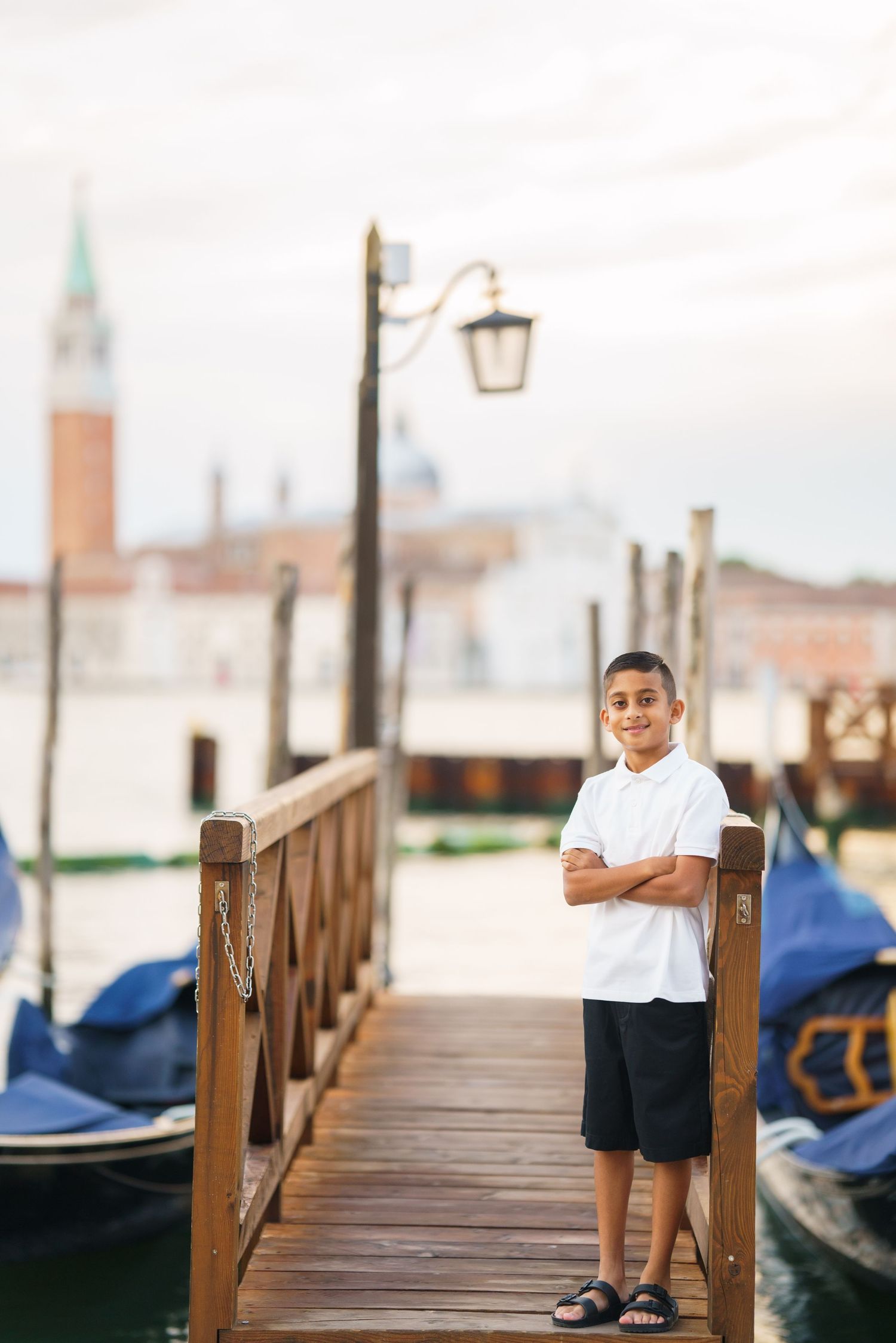 A boy with crossed arms stands on a wooden dock in Venice, Italy, with gondolas and San Giorgio Maggiore in the background.