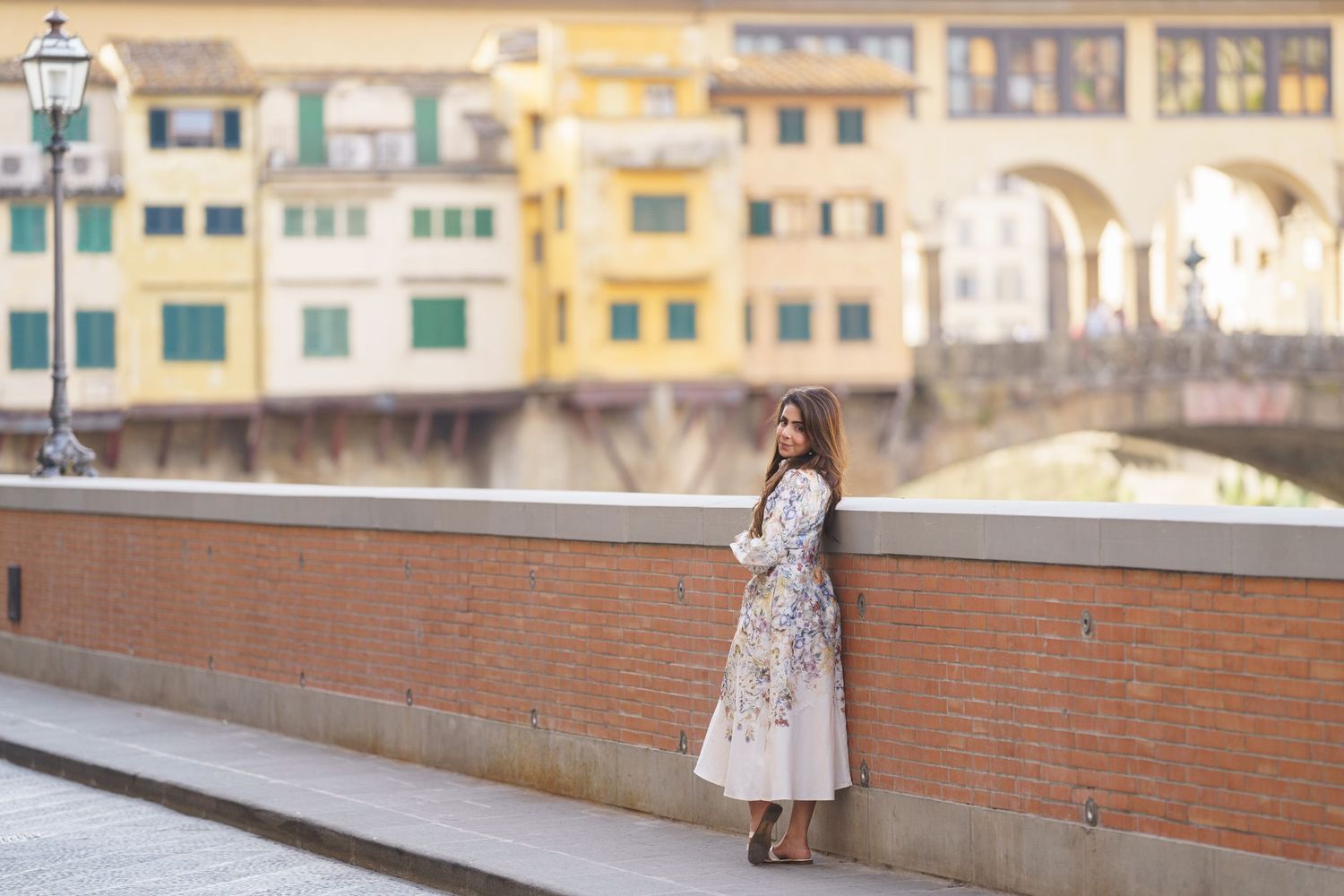 Woman in floral dress poses by brick wall with Ponte Vecchio bridge in Florence, Italy in background.