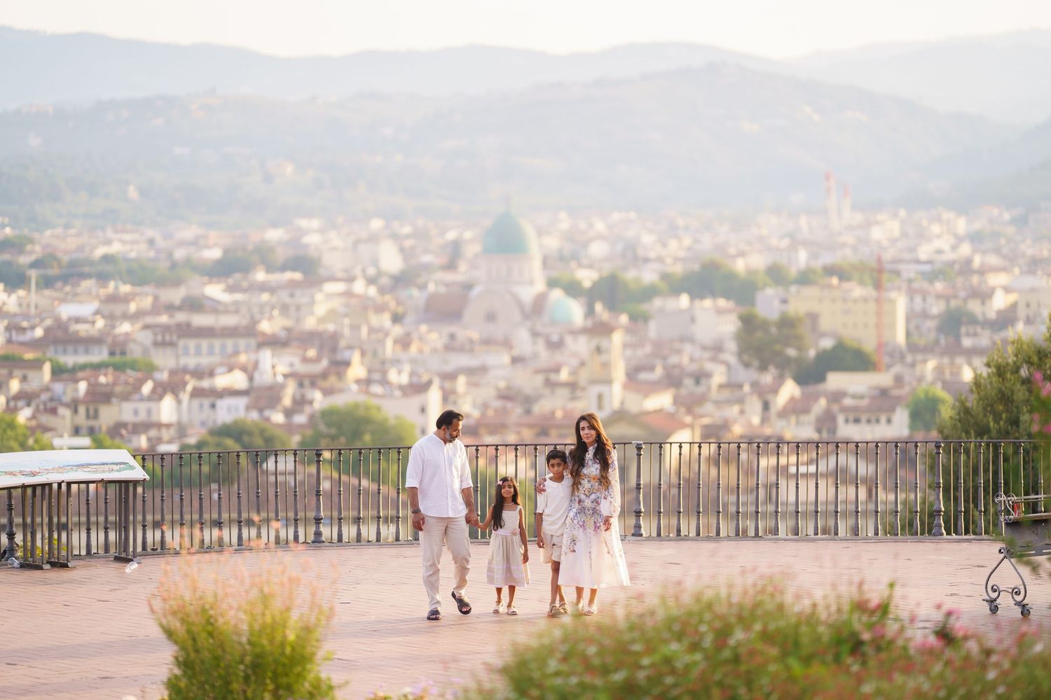 Family of four in white outfits strolling on a terrace overlooking Florence cityscape with cathedral dome at sunset.