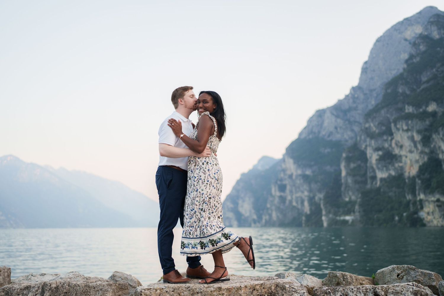Couple sharing a romantic kiss on rocky lakeside with dramatic mountain cliffs and serene water backdrop.
