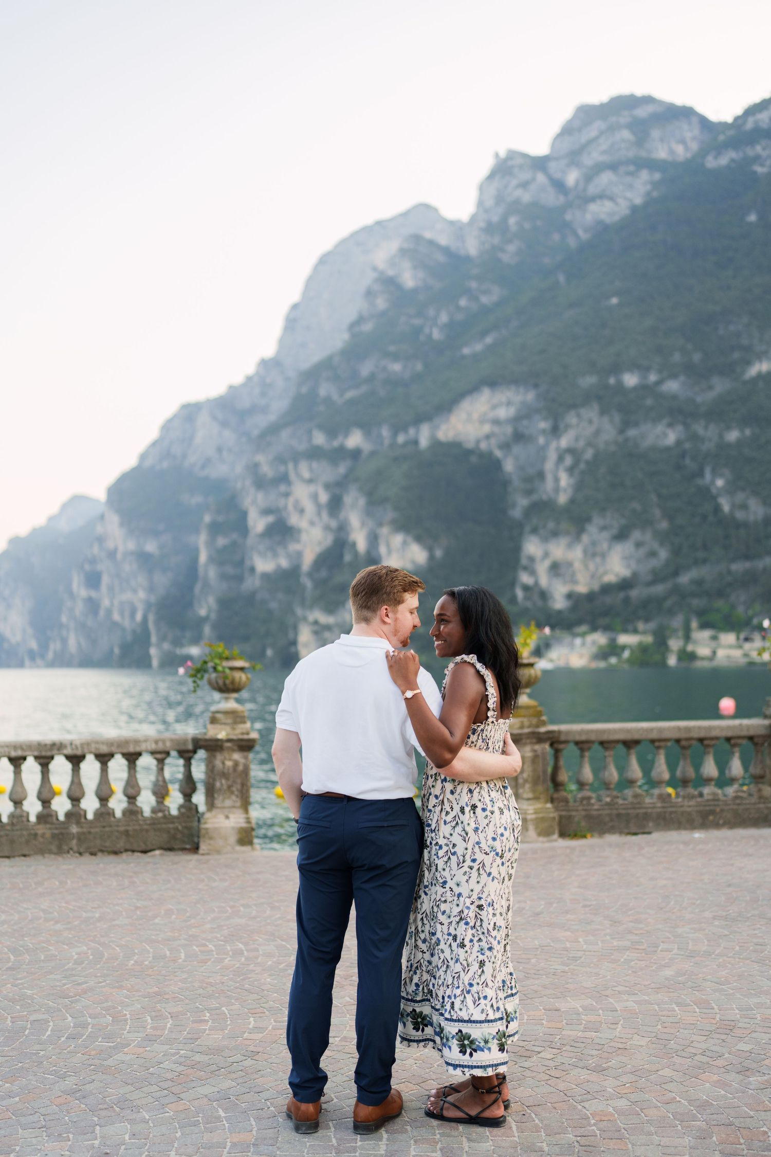Romantic interracial couple embracing on a lakeside terrace with dramatic mountain backdrop.