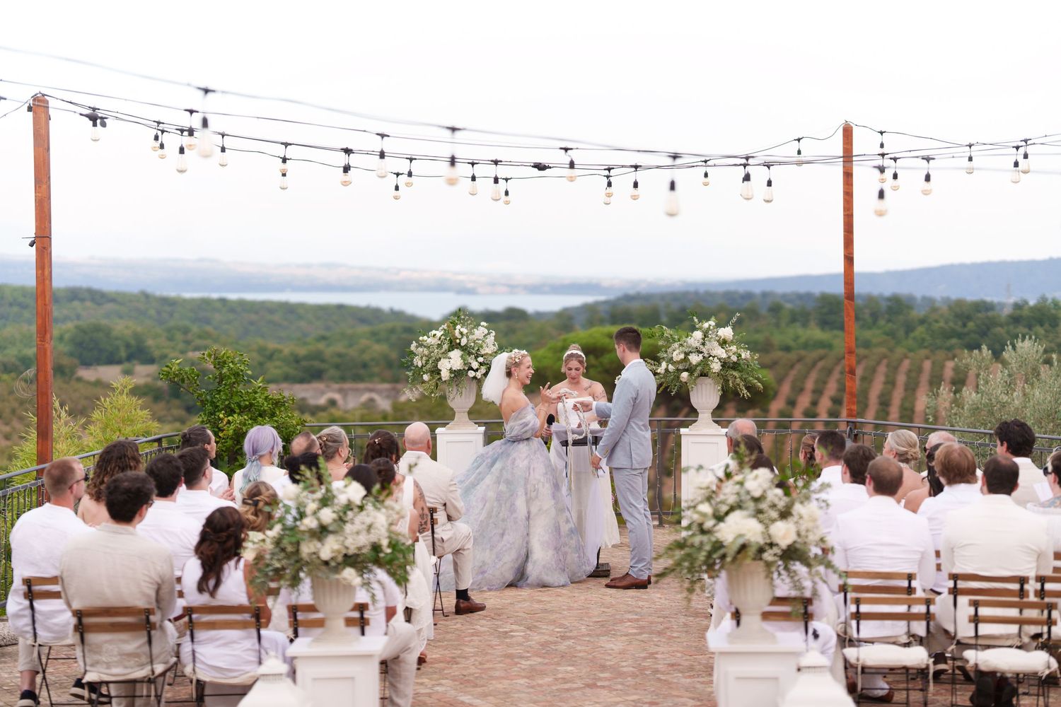 Outdoor wedding ceremony on a terrace with string lights, white floral arrangements, and scenic valley views.