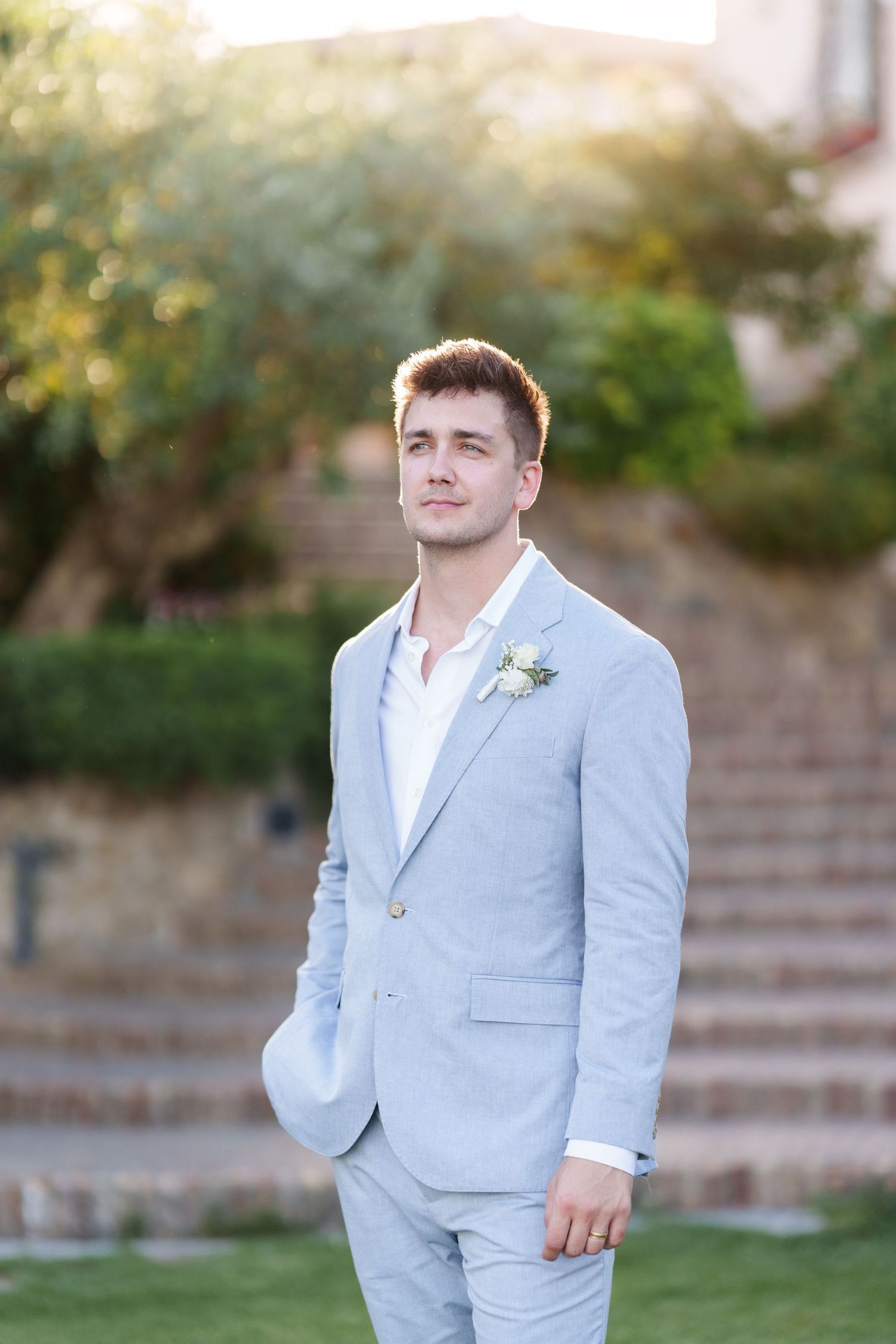 Young groom in light blue suit with boutonniere poses outdoors on green lawn with stone steps and trees in background.