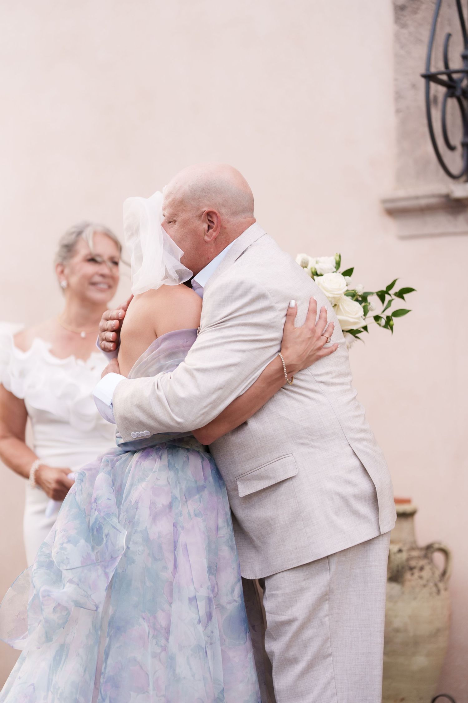 Bride in floral gown hugging bald groom in gray suit at outdoor wedding ceremony with white rose bouquet.