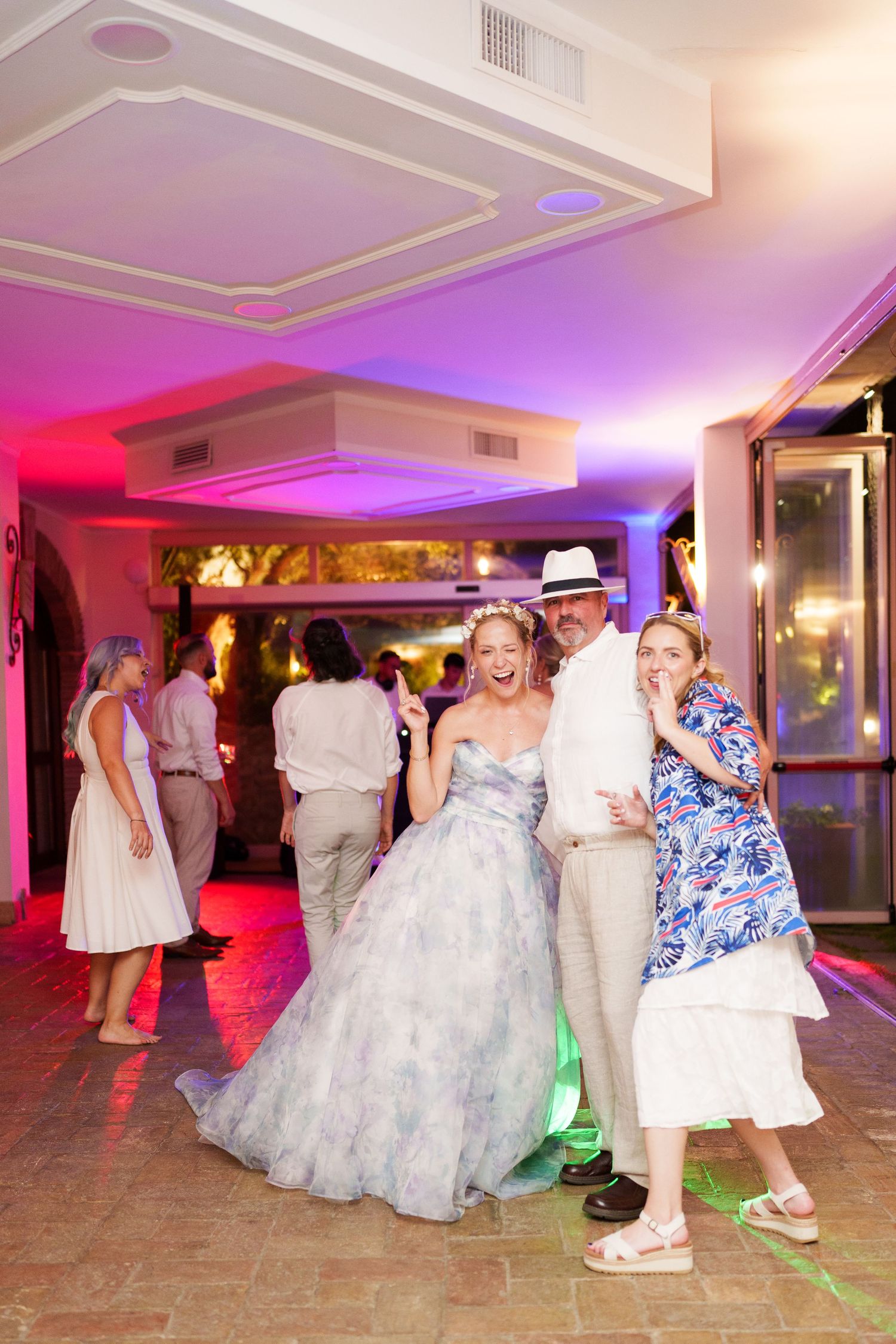 Bride in floral gown poses with guests on wedding reception dance floor with colorful purple and pink lighting.