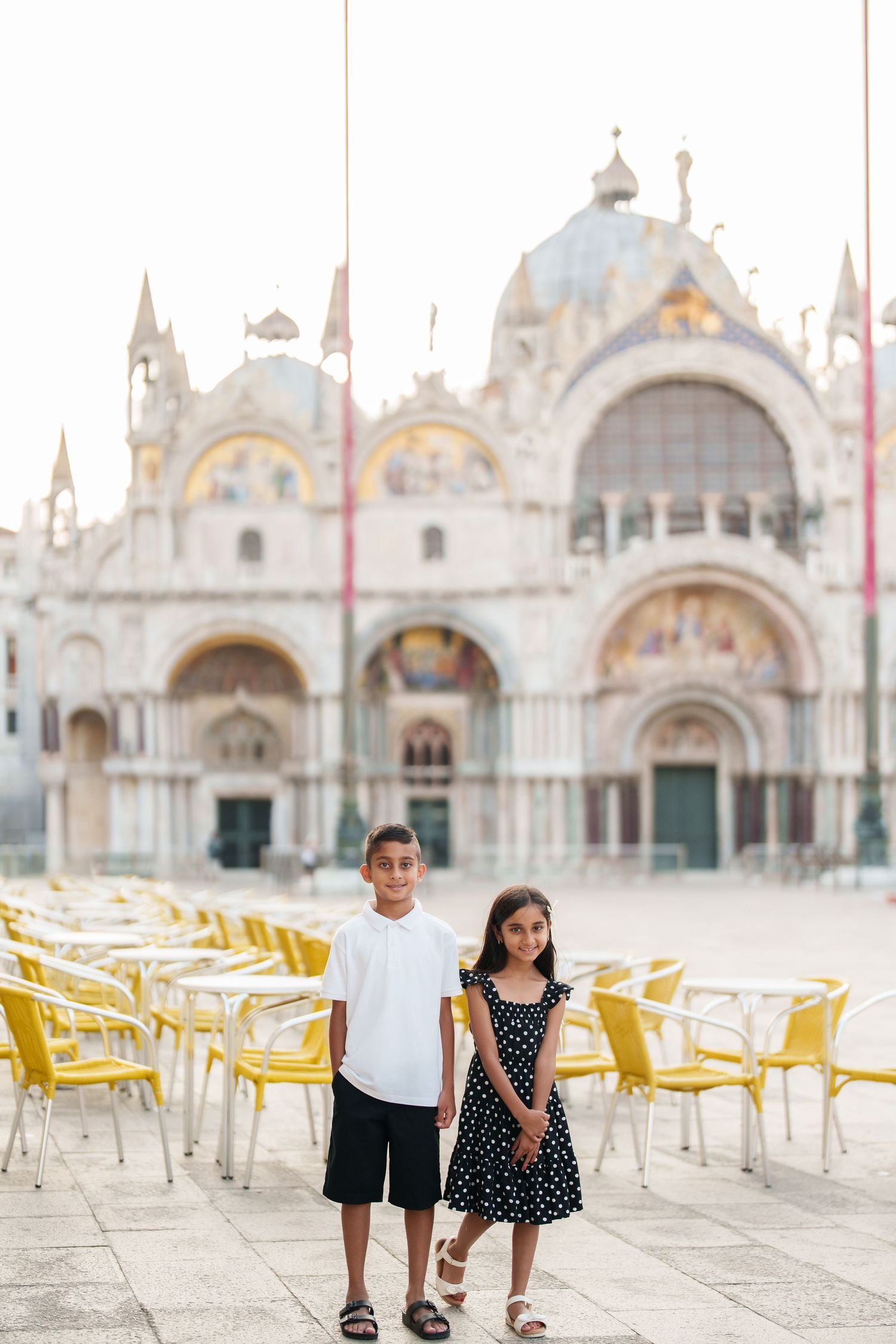 Two children posing in St. Mark's Square, Venice, with the iconic Basilica di San Marco in the background.