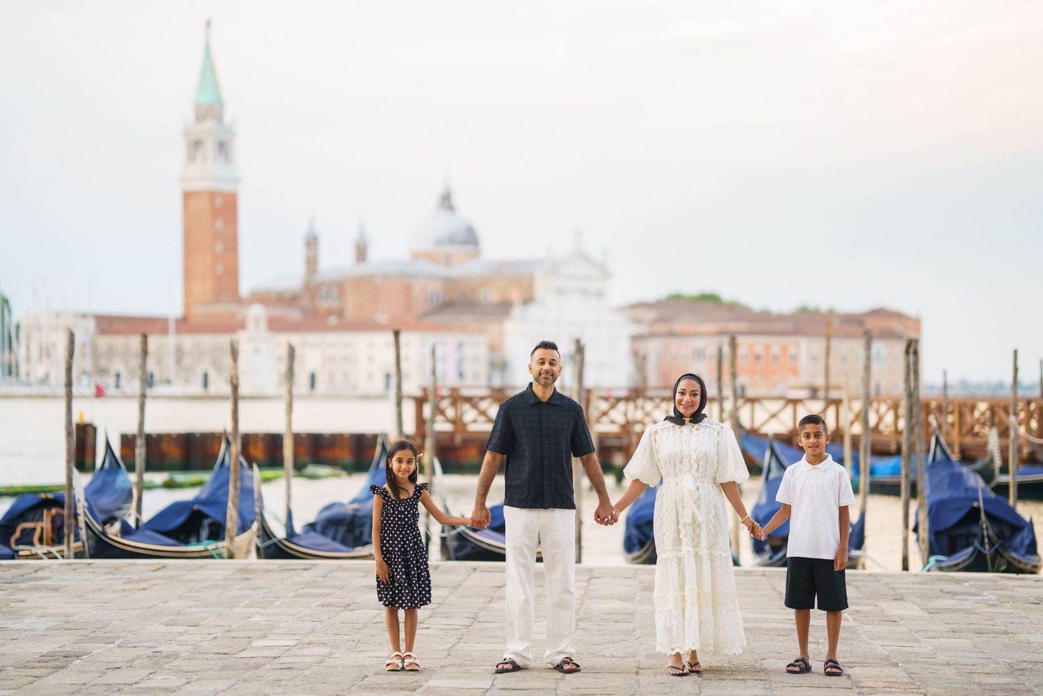 Family of four holding hands on Venice waterfront with gondolas and San Giorgio Maggiore church in background.