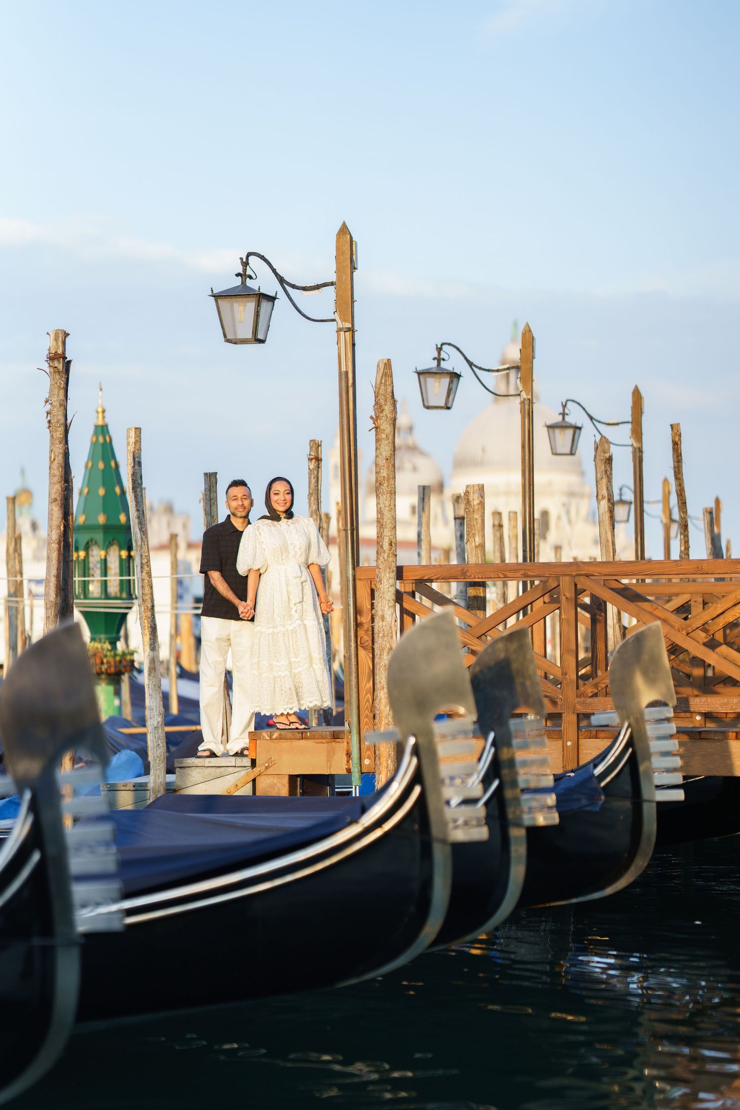 Couple standing on a gondola dock in Venice, Italy, with the Santa Maria della Salute basilica in the background.
