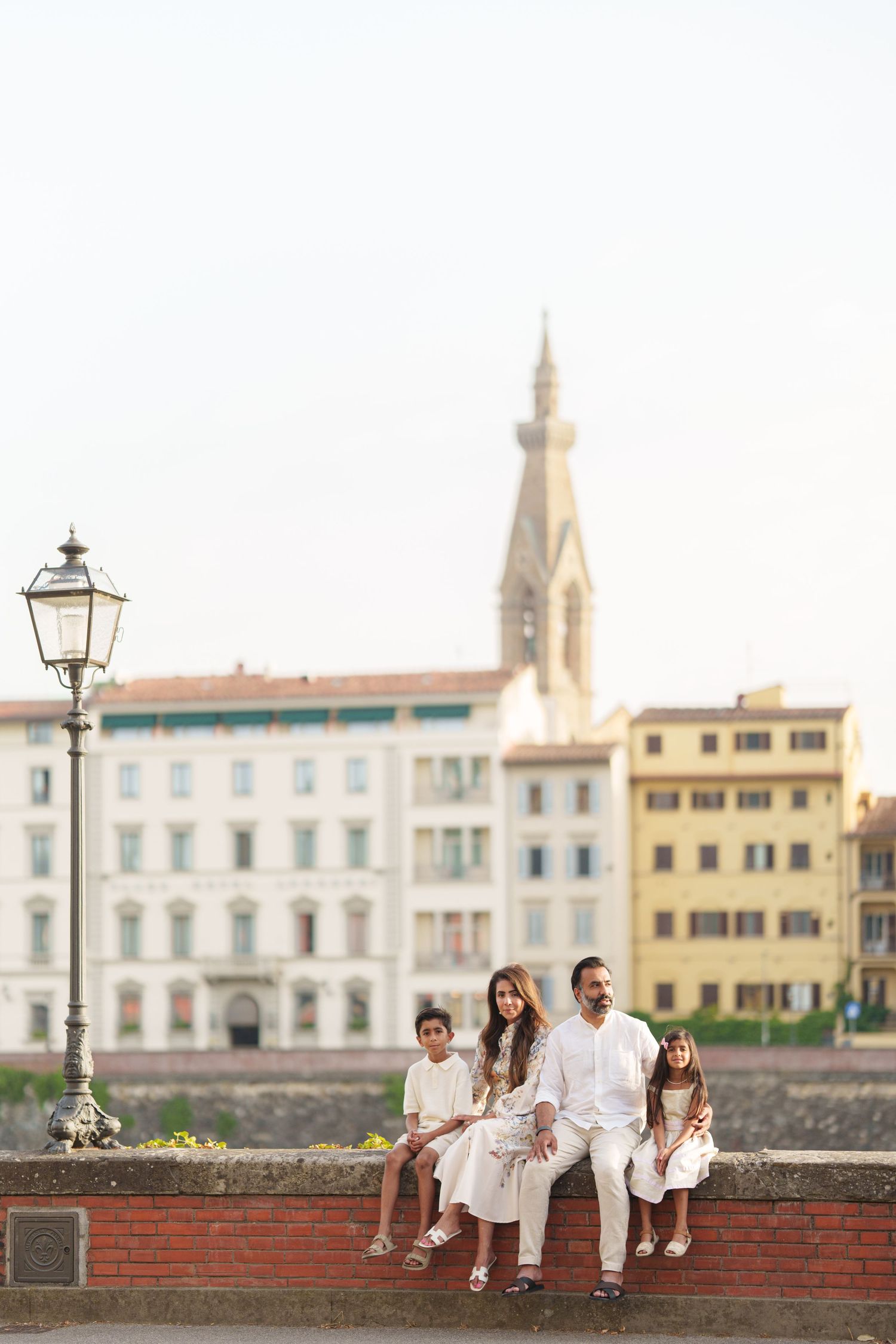 Family of four in white outfits sitting on a bridge wall in Florence, Italy, with historic buildings and church tower behind.