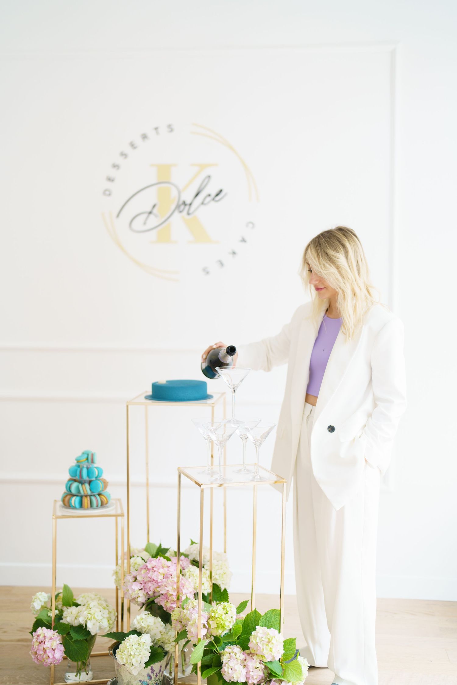 Blonde woman in white suit pouring champagne at elegant dessert display with blue macarons and floral arrangements.