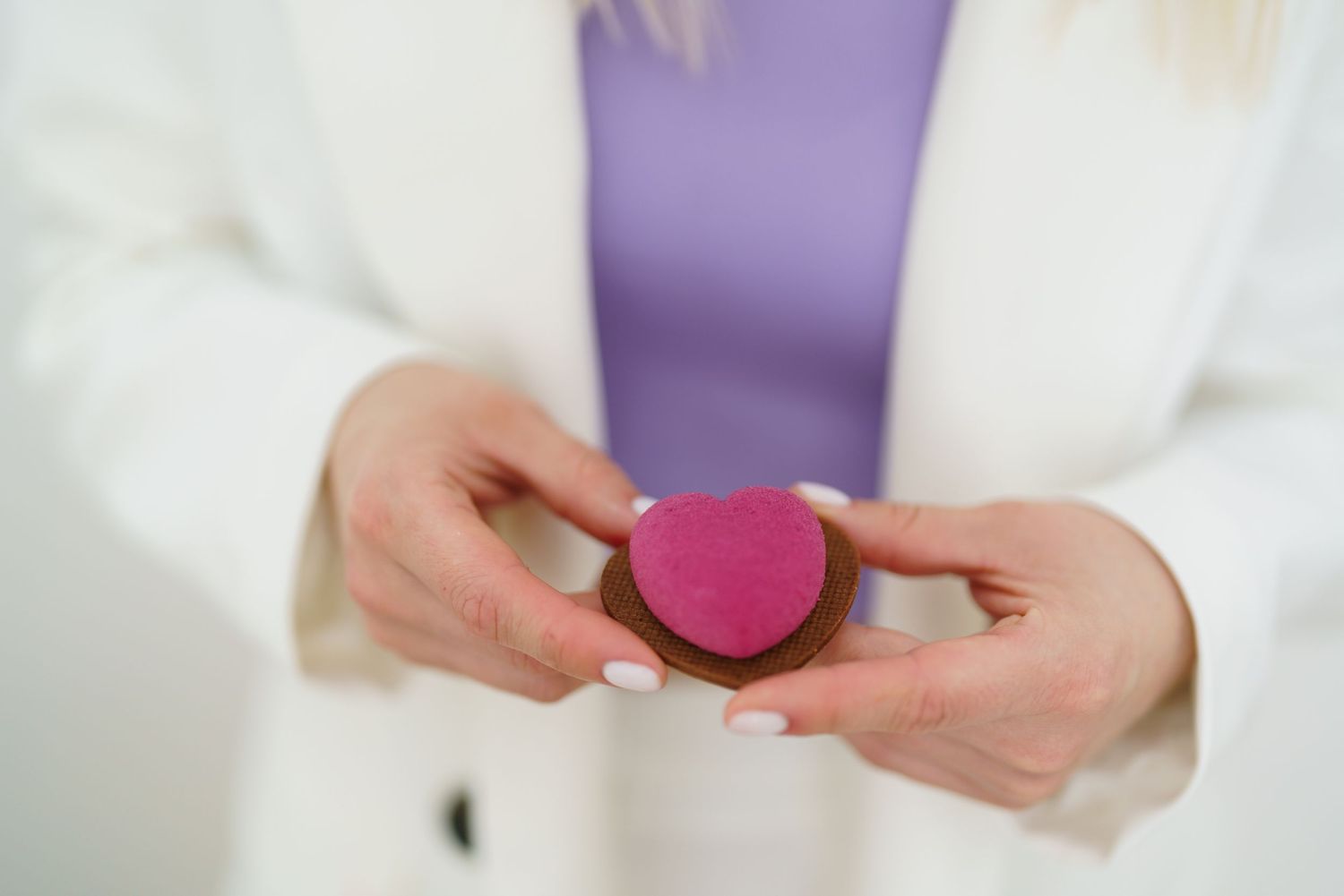 Woman in white blazer holding a pink heart-shaped chocolate macaron with both hands.