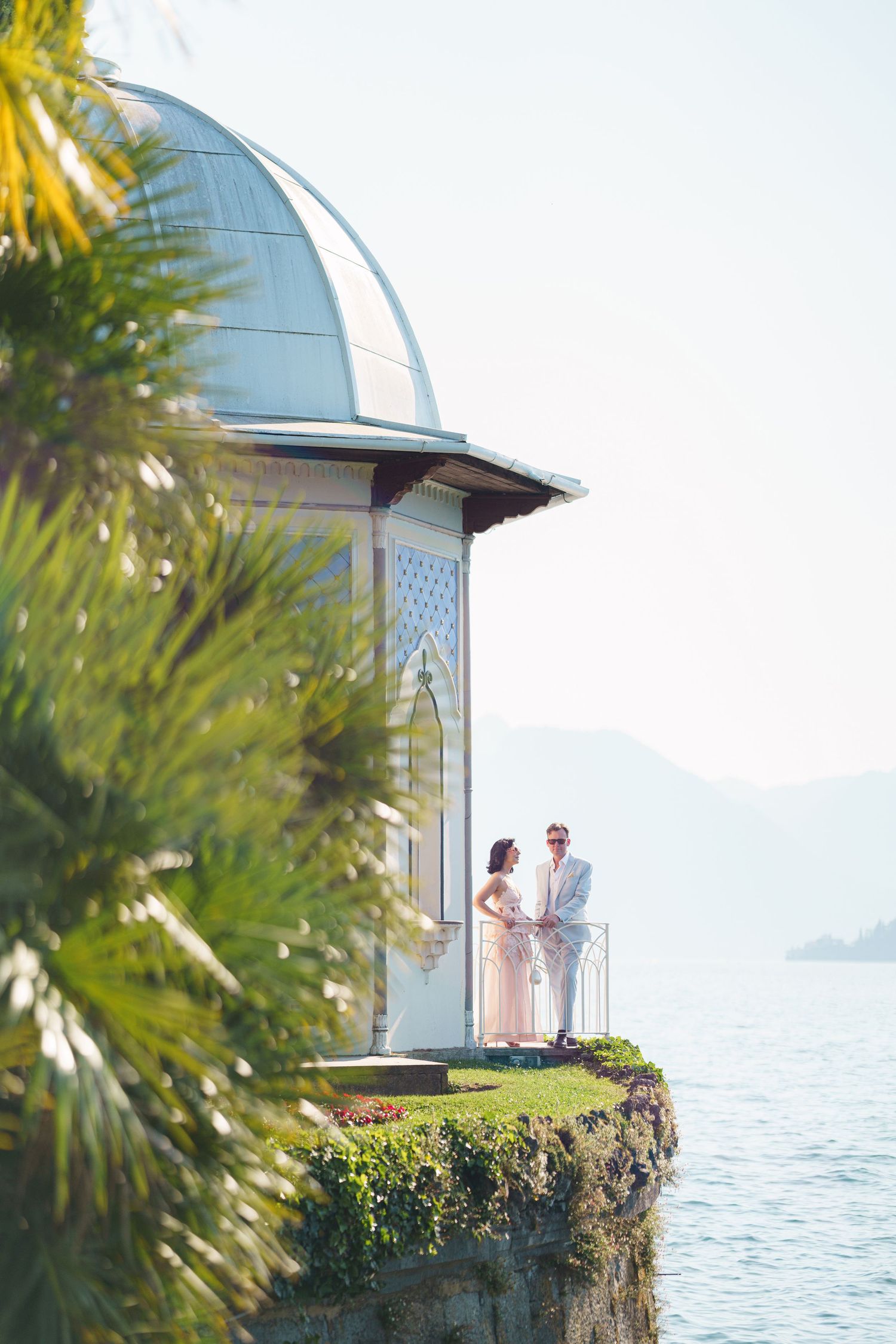 Couple standing on elegant lakeside pavilion balcony with palm trees and mountain views on a sunny day.
