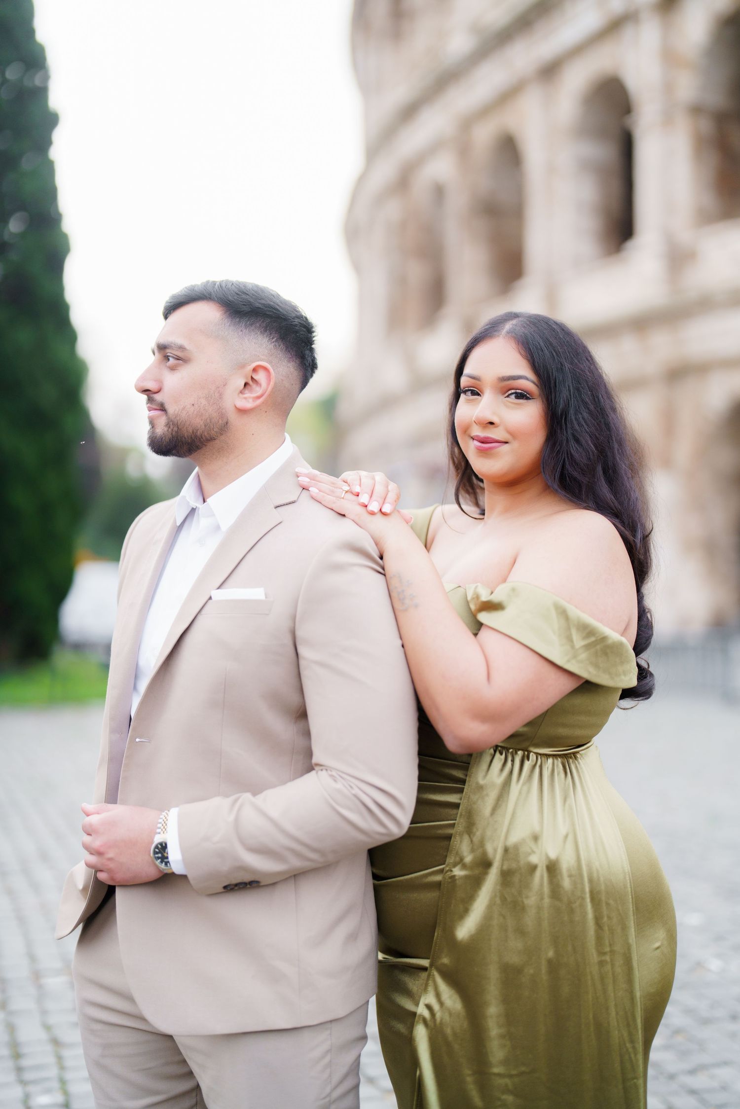 Couple posing near the Colosseum in Rome, man in beige suit and woman in elegant olive green off-shoulder gown.