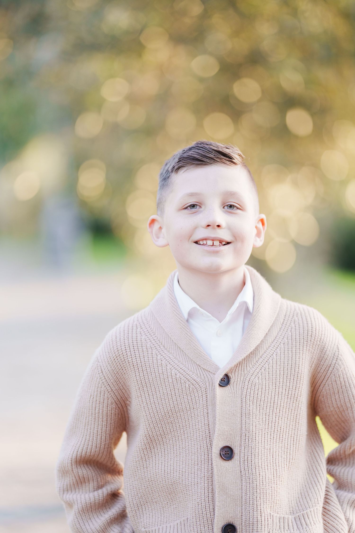 A smiling young boy in a beige cardigan and white shirt poses outdoors with a soft bokeh background.