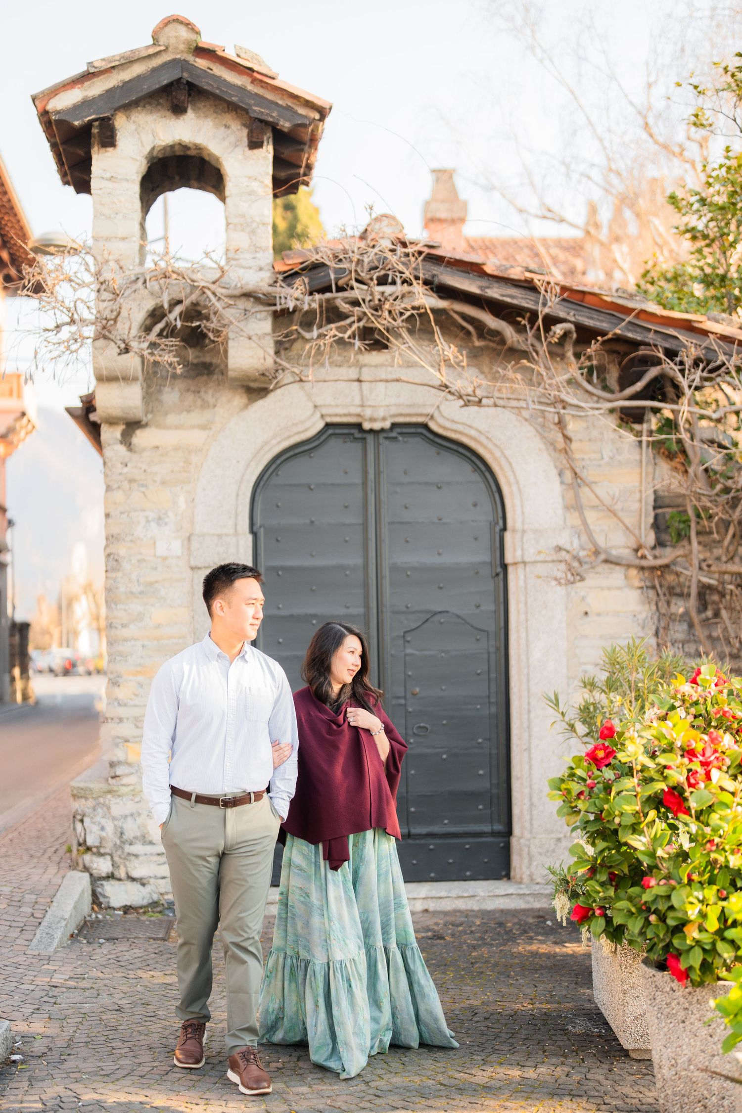 Couple posing in front of a stone archway with vintage wooden doors and red flowers in a European village.