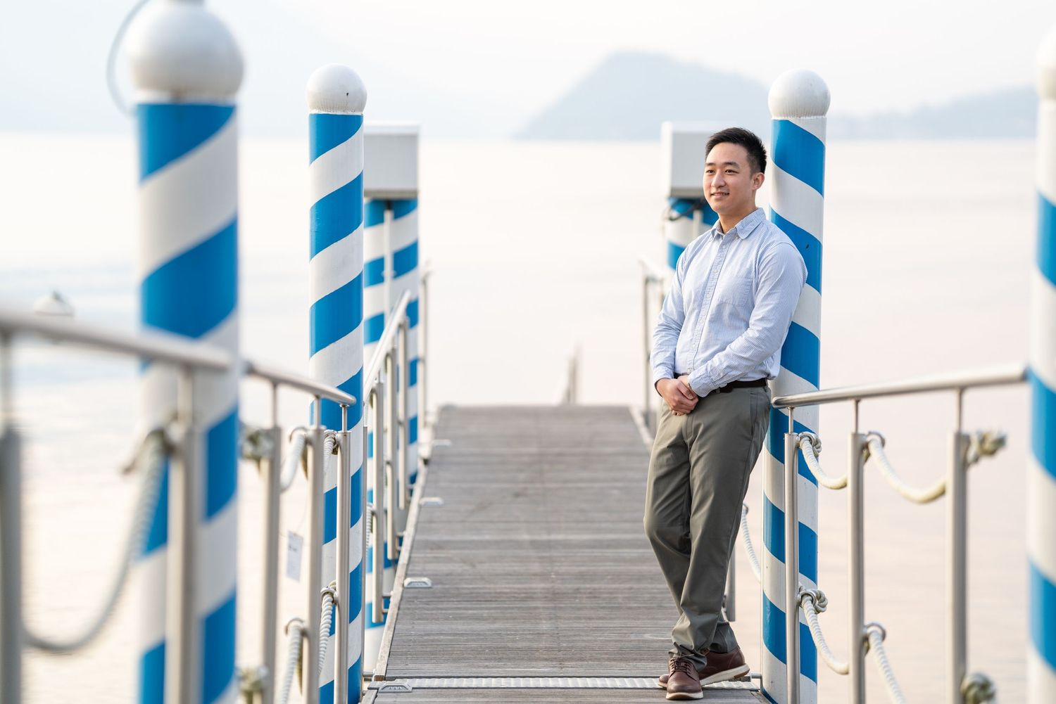 Asian man in light blue shirt leans on railing of blue and white striped pier overlooking calm ocean.