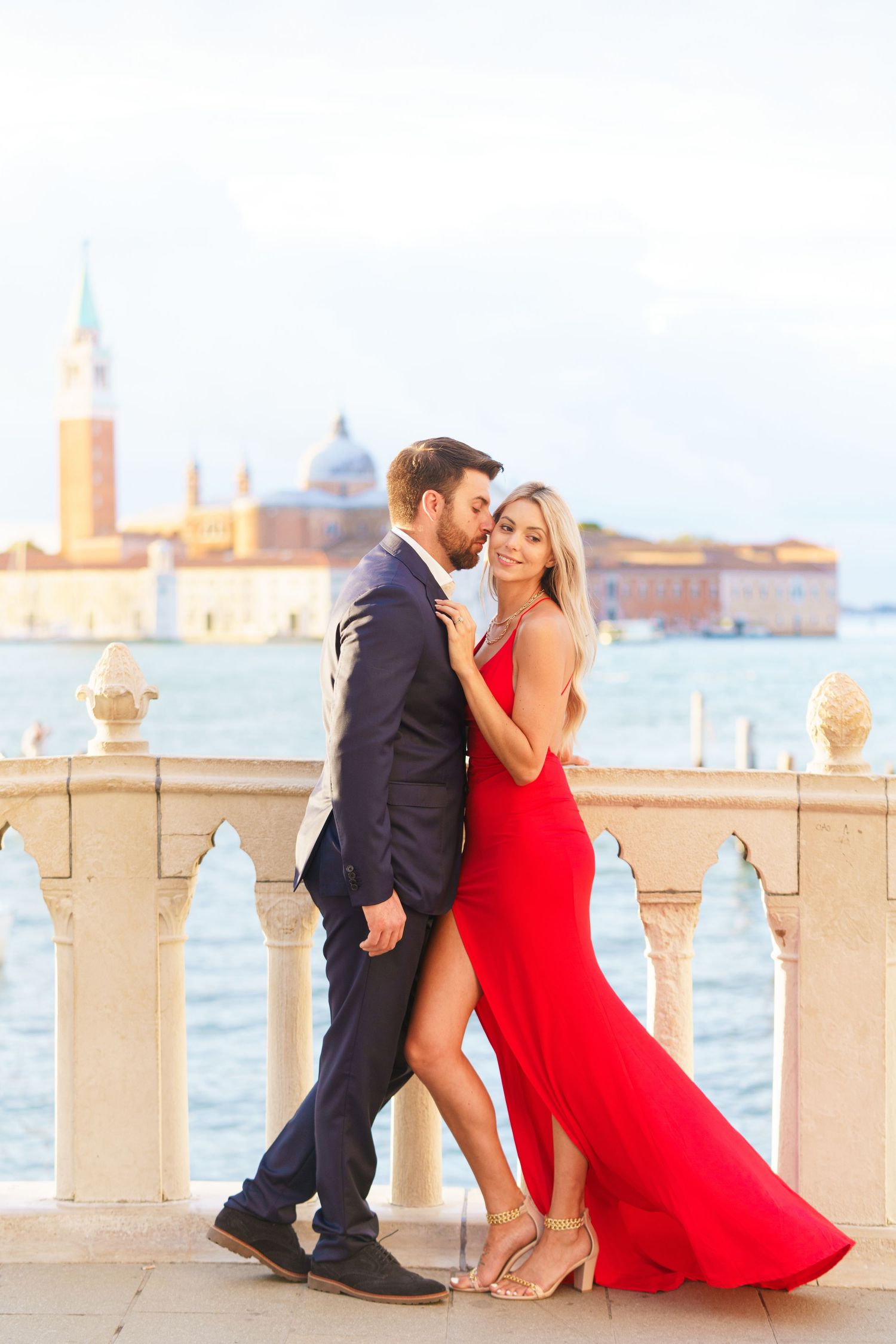 A couple in formal attire shares romantic moments along a waterfront balustrade in Venice at sunset.