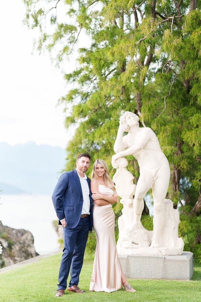 A couple poses near white stone sculptures in a garden setting with green trees in the background.