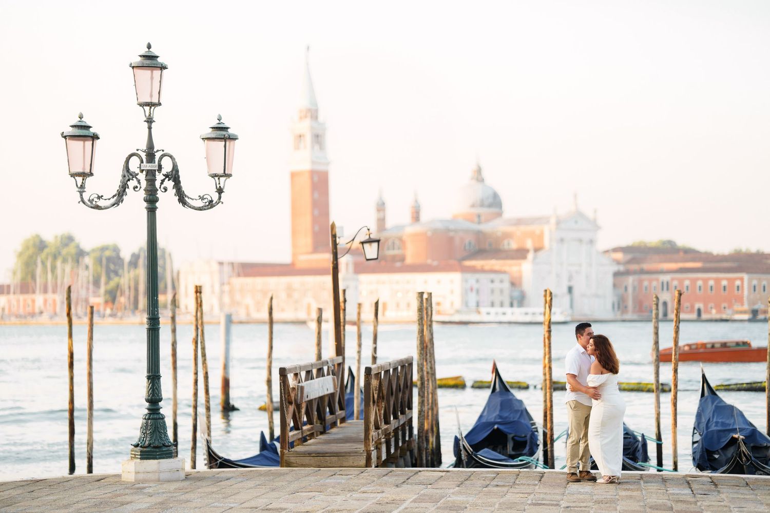 Panoramic view of Venice waterfront with gondolas, ornate lampposts, and the iconic San Giorgio Maggiore church in the distance.