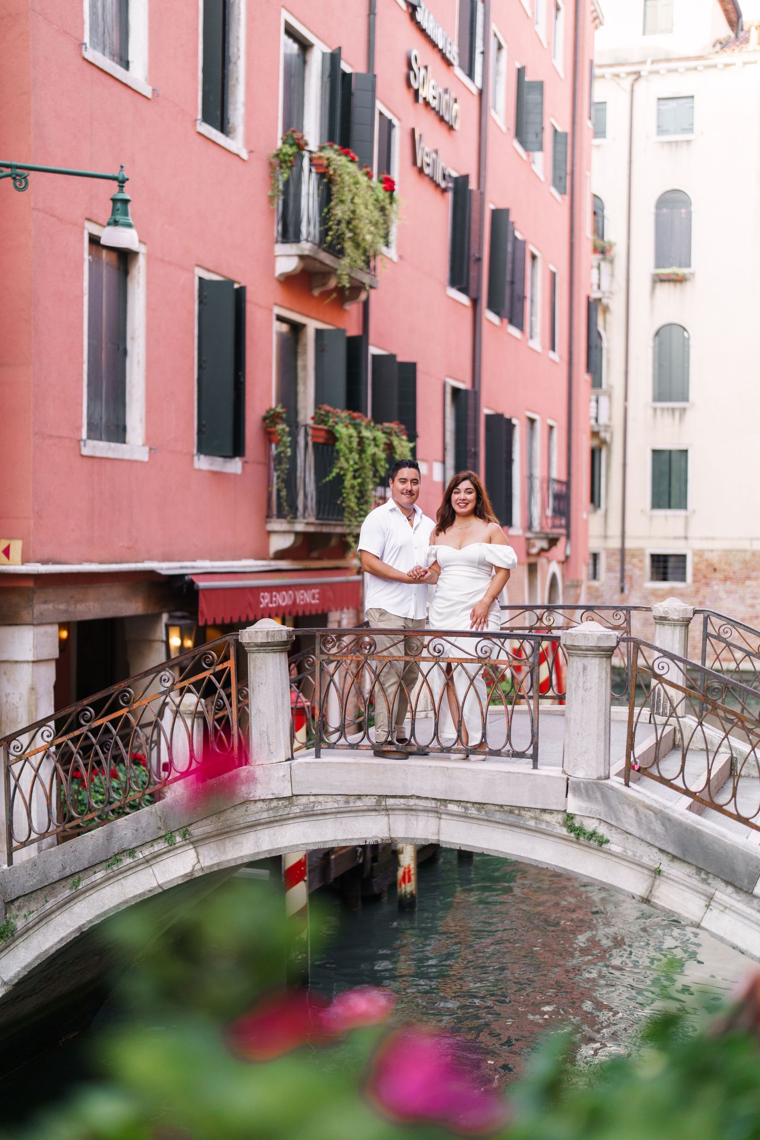 Romantic couple standing on a small arched bridge over a Venice canal with pink buildings and flowers in the foreground.