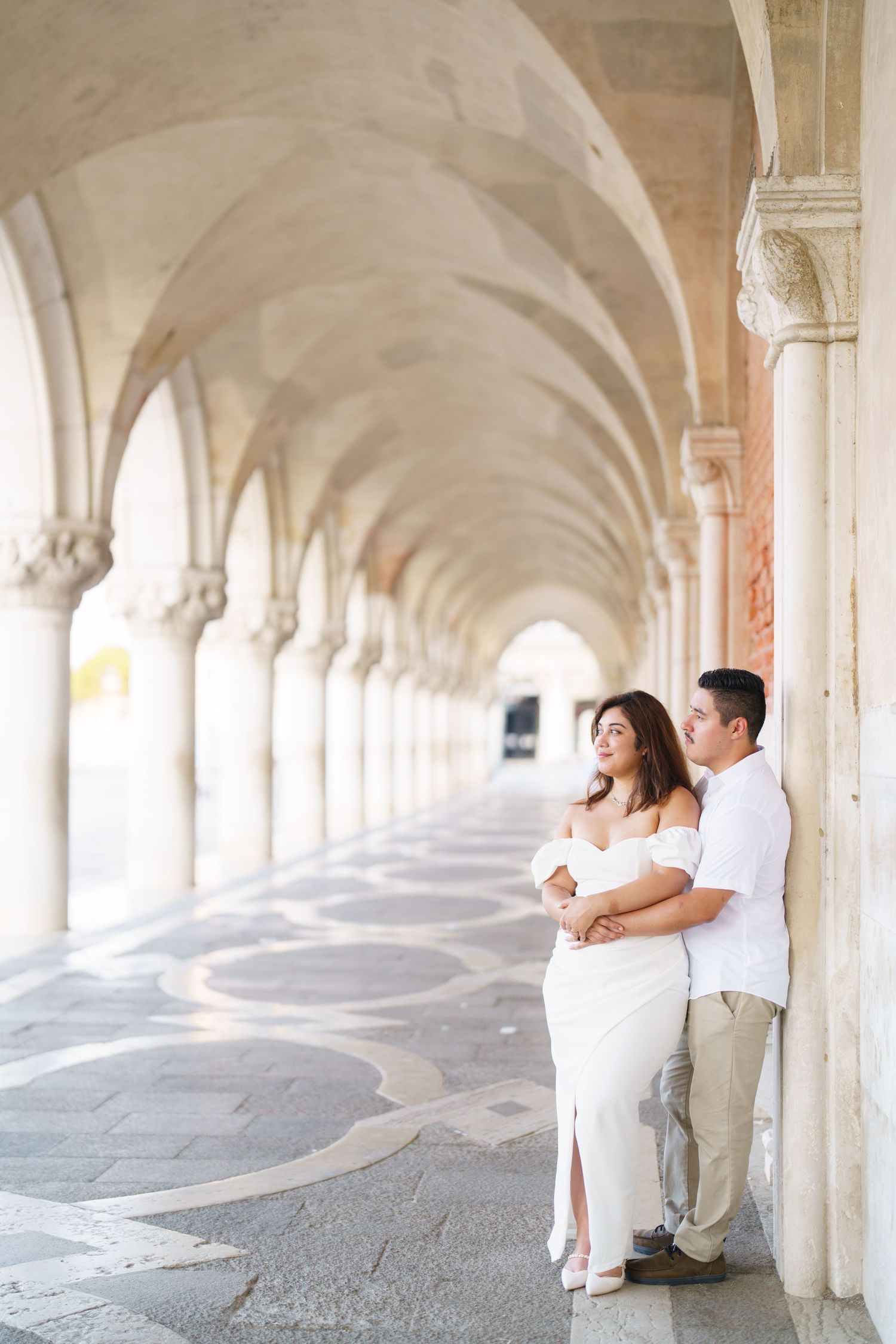 A couple in white outfits embraces under elegant arched stone colonnade during engagement photoshoot.