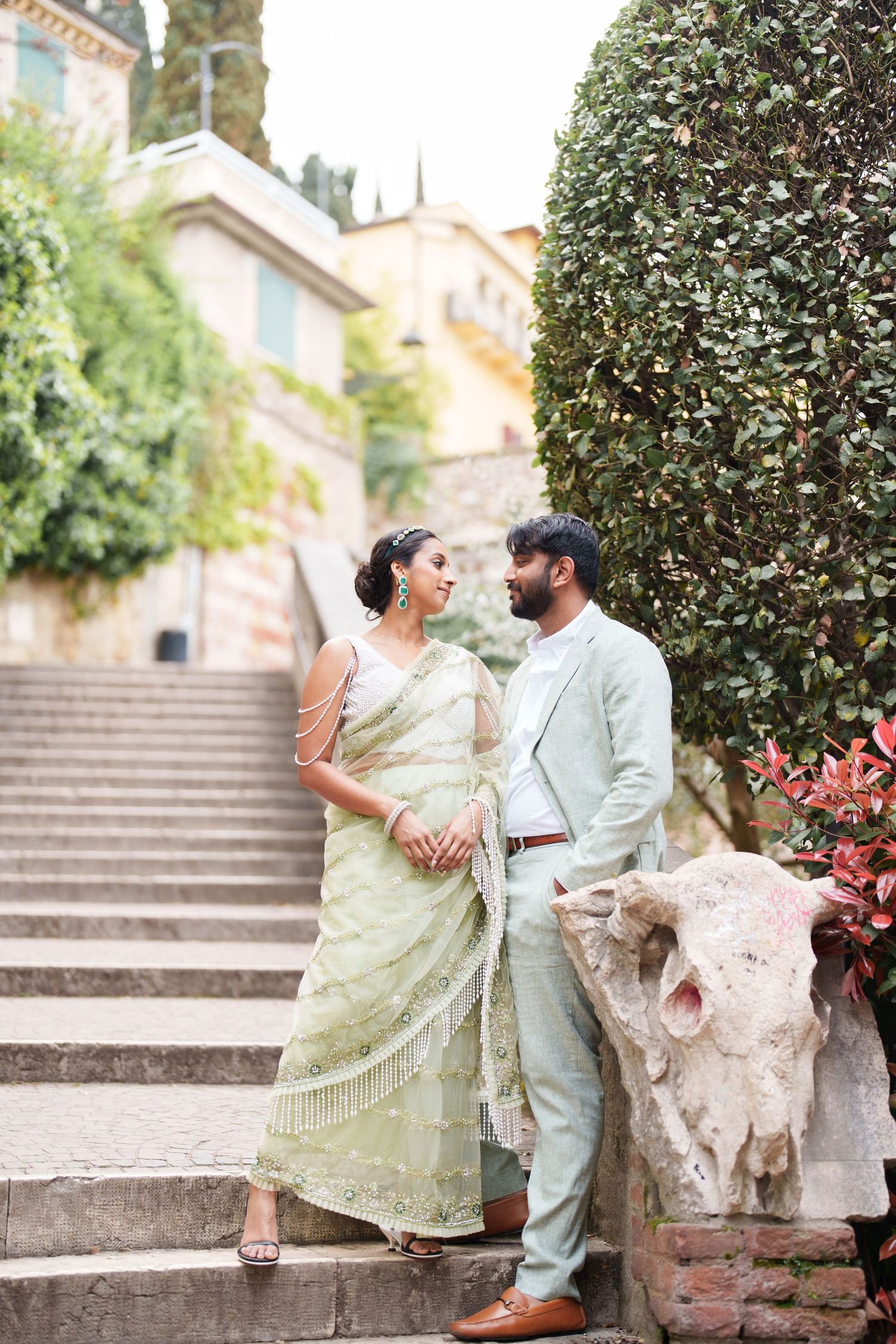A romantic couple in traditional Indian attire shares intimate moments on stone steps surrounded by greenery and flowers.