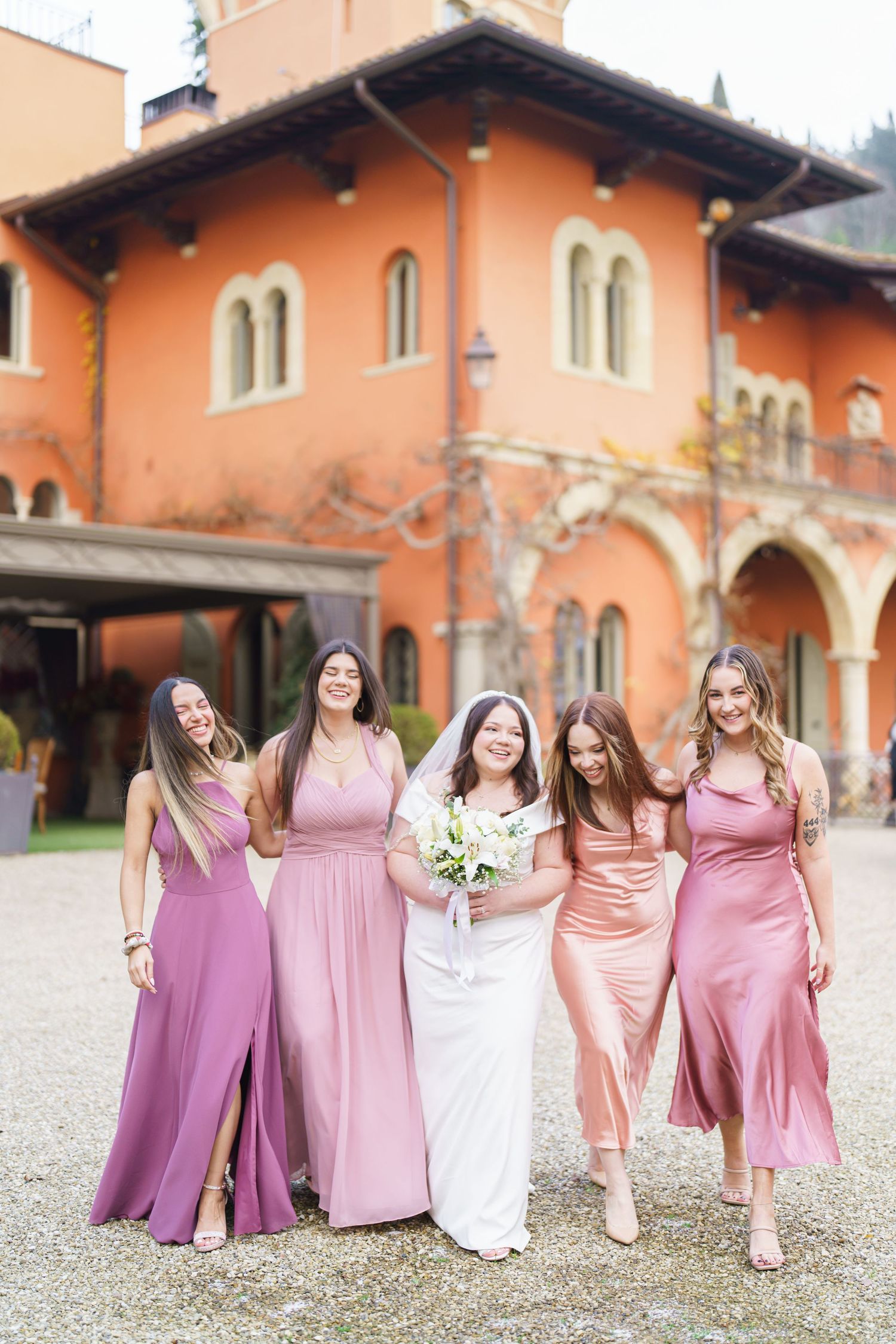 Wedding party poses in pink and purple bridesmaid dresses outside a Spanish-style building.