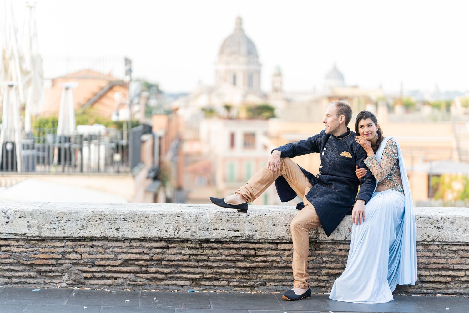 Couple in fusion wedding attire sitting on stone wall with Rome cityscape in background.