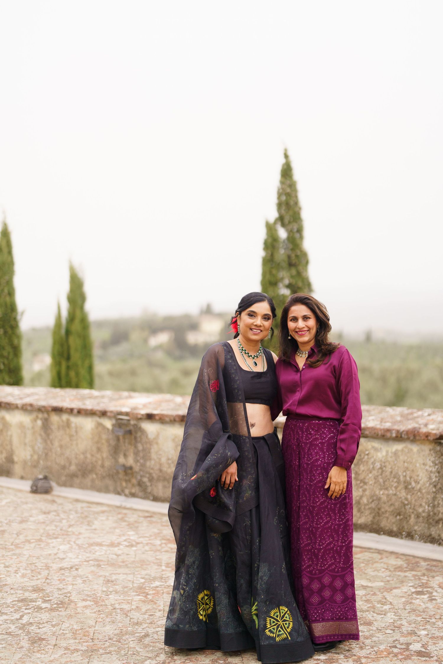 Two people in dark and burgundy attire pose together against a stone wall with cypress trees.