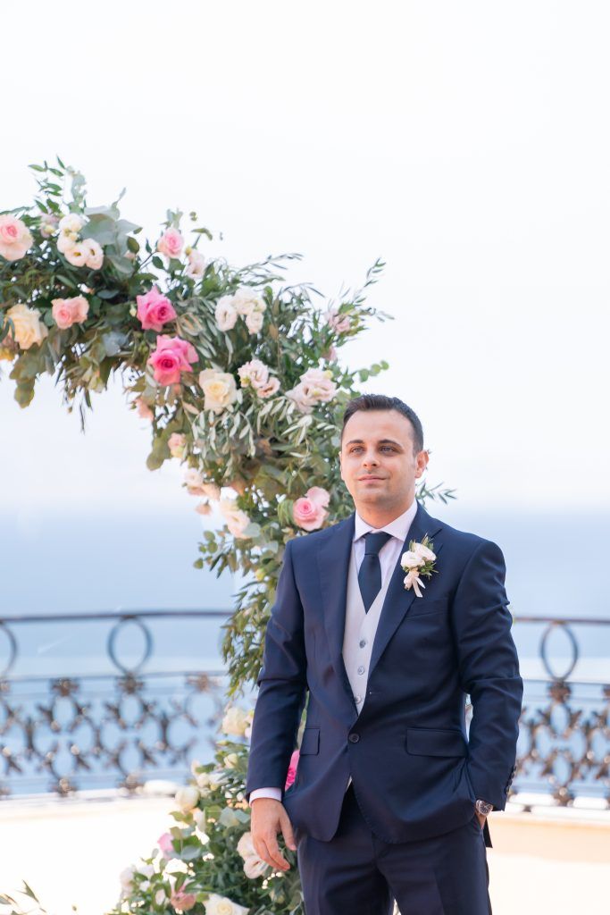 Groomsmen in navy suits stand outdoors against a scenic backdrop with pink floral arrangements.