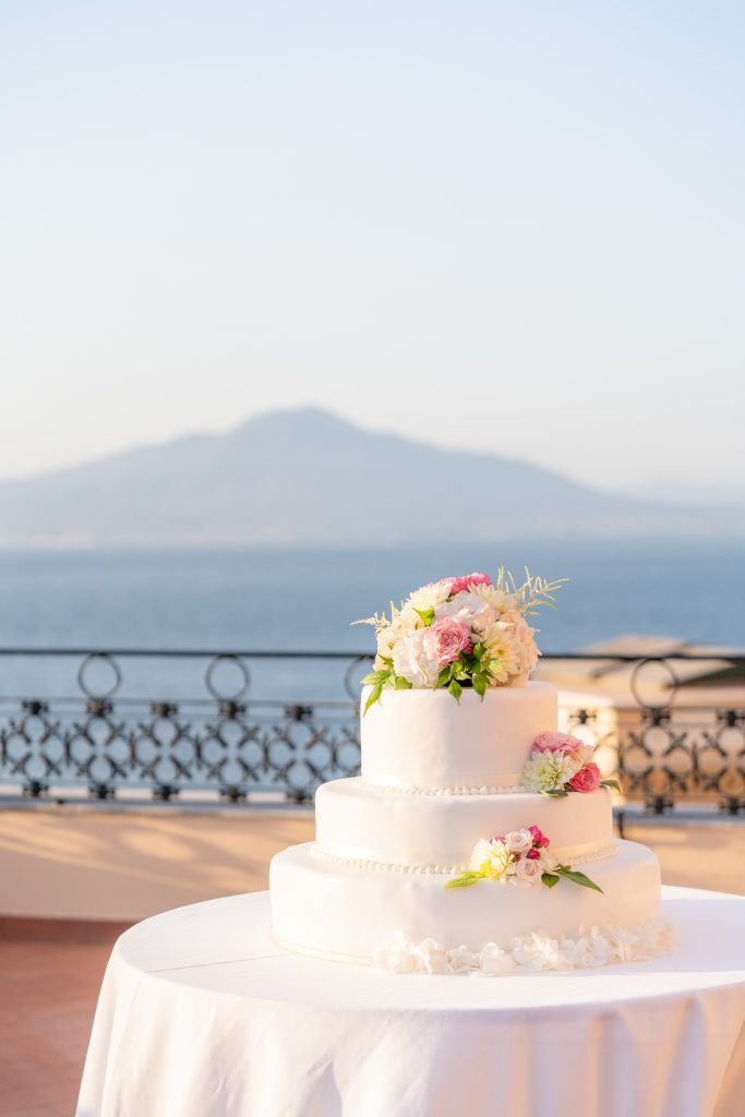 White wedding cake decorated with pink flowers on a terrace with coastal views at sunset.