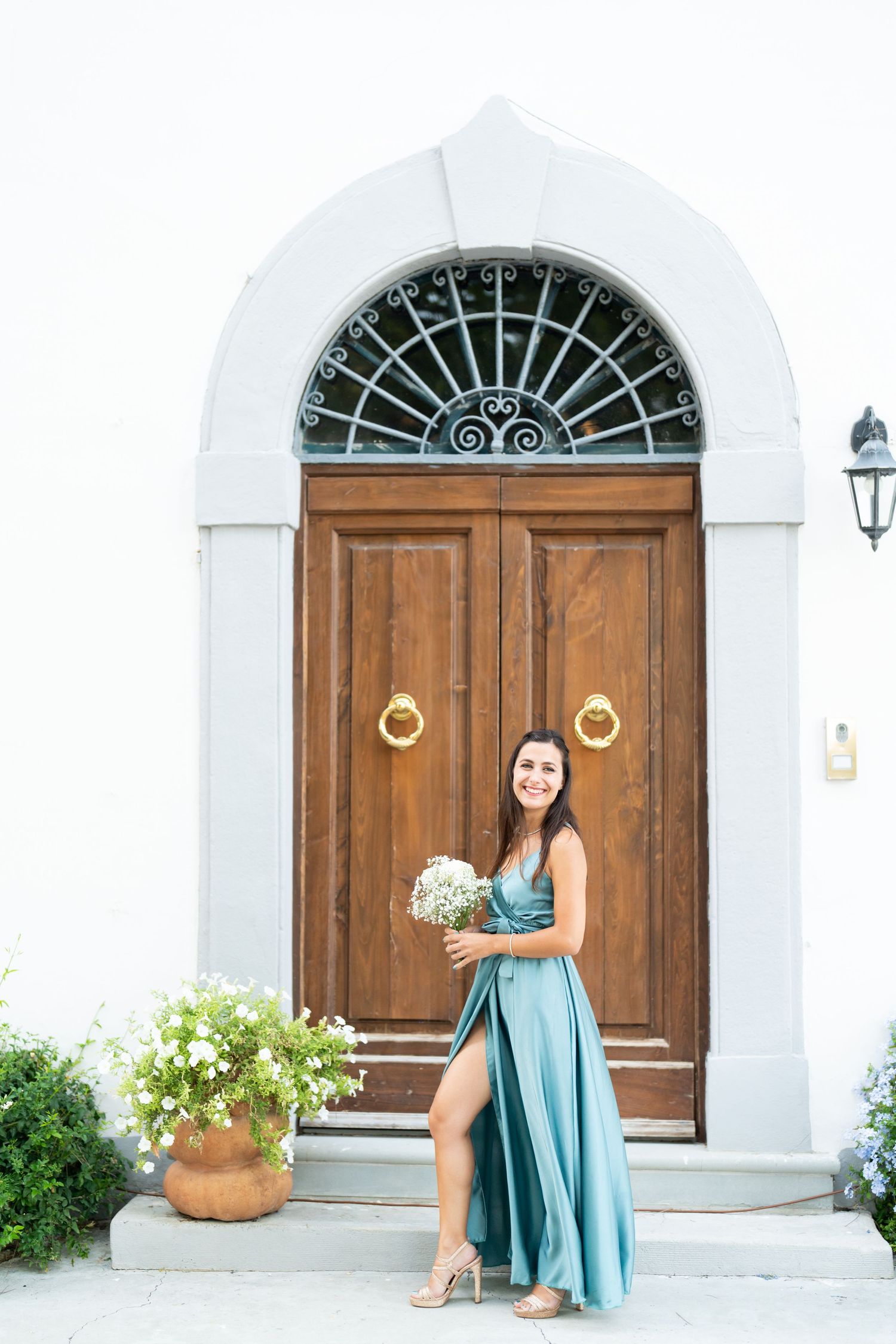 A bridesmaid in a teal gown poses with flowers by an elegant arched wooden door.