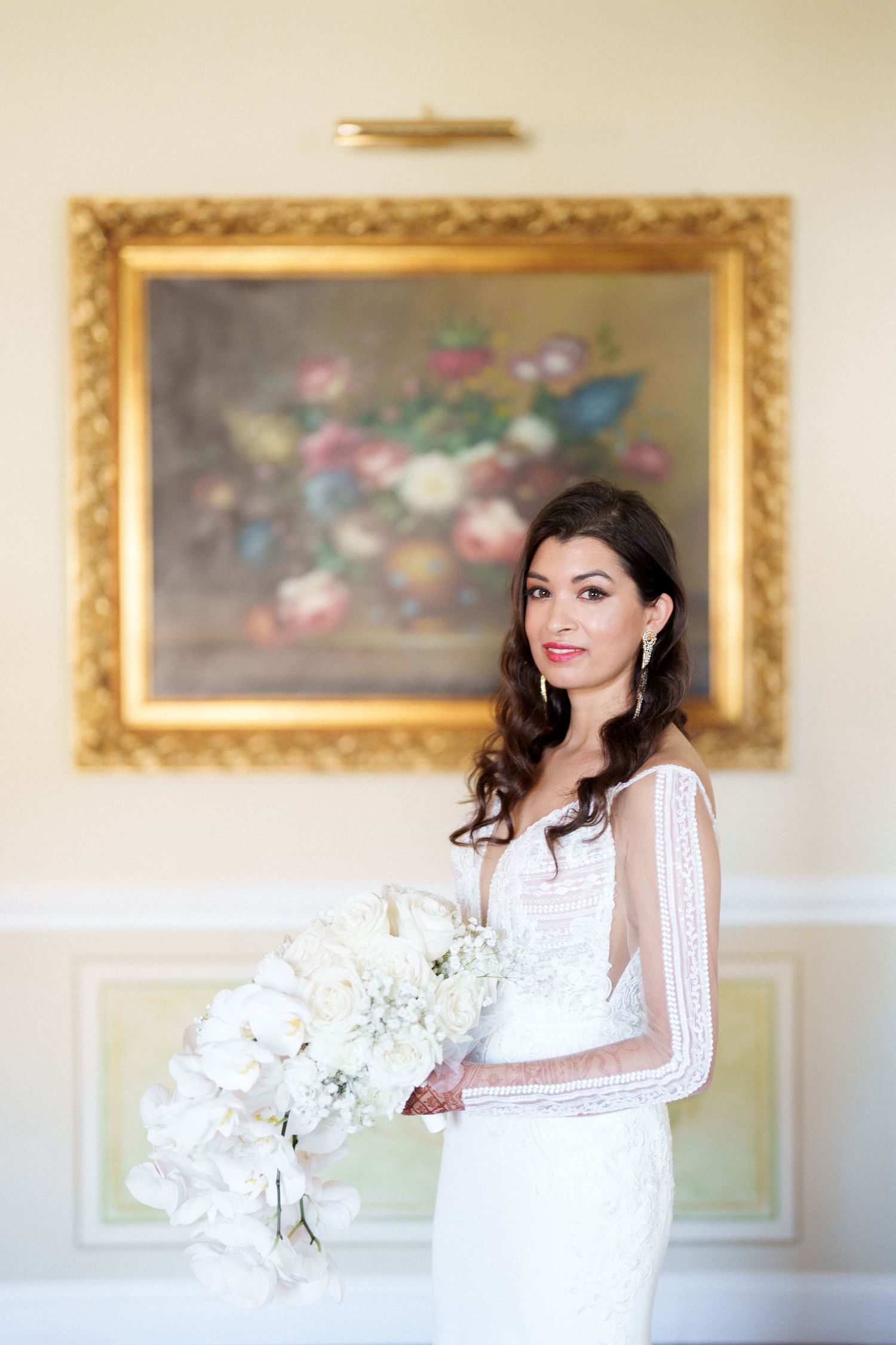 A person in white lace holds delicate white flowers against an elegant wall with framed artwork.